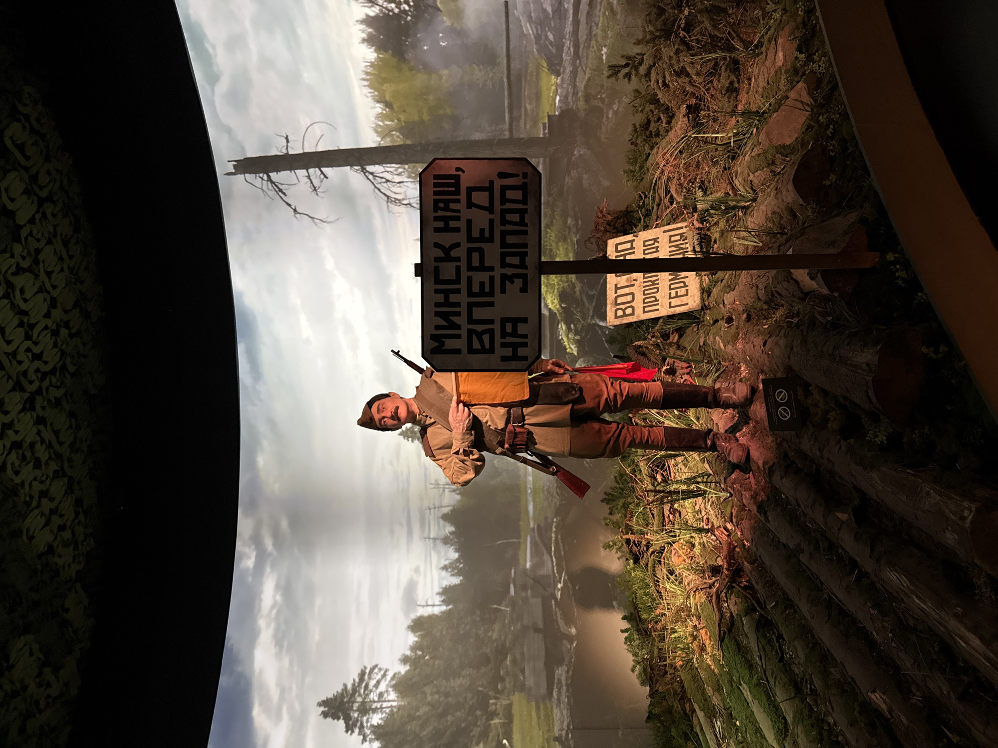 This media file depicts a diorama set in Zelenaya Roshcha, Russia. The scene features a solitary male figure dressed in a military uniform, appearing to be from the mid-20th century. He is wearing a cap, a uniform jacket with a bandolier, trousers, and boots. He holds a rifle and stands on a log-strewn embankment, with a backdrop of a misty forest and cloudy sky.
Two signs are prominently displayed in the scene. The larger sign, positioned behind the soldier, is black with white lettering in Russian that reads: "МИНСК НАШ, ВПЕРЕД НА ЗАПАД!" This translates to "Minsk is ours, forward to the West!" The smaller sign, to the right of the first, also in Russian, is partially visible and reads: "ВОТ, АД ПРОКЛЯТИЯ ГЕРМАНИИ!" meaning "Here is the hell of Germany's damnation!" These inscriptions suggest a wartime setting, likely related to the Eastern Front during World War II.
The lighting suggests an indoor display, possibly a museum exhibit, designed to evoke a specific historical moment. The overall mood is somber and evocative of conflict. The diorama's realistic portrayal of the landscape, including trees, moss, and rough terrain, contributes to the immersive quality of the scene. The backdrop painting creates a sense of depth and distance, further enhancing the illusion of a real environment.
