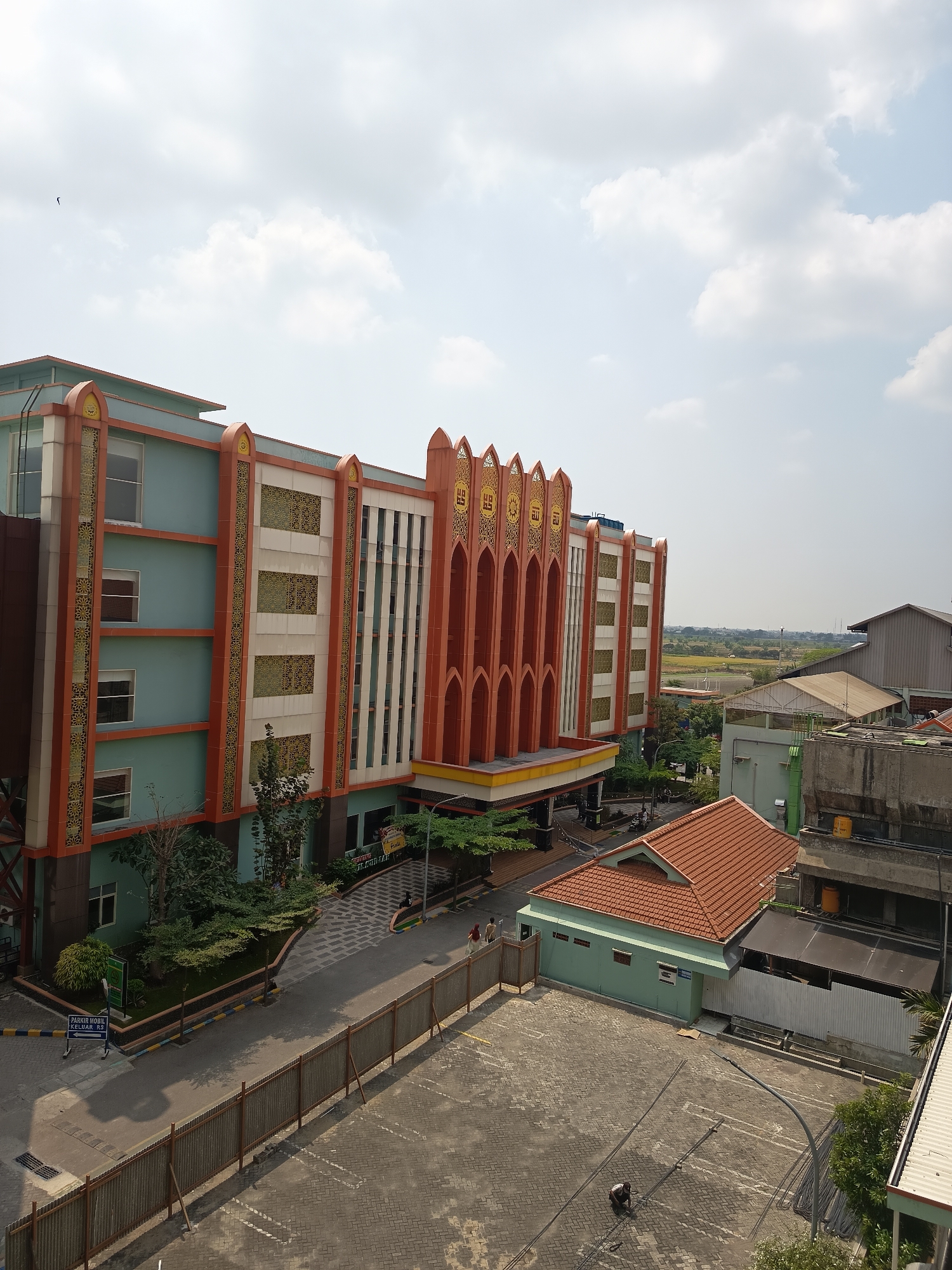 This is an aerial, eye-level photograph taken on a clear day in Dapur, Indonesia. The scene is dominated by a multi-story building with a distinctive, ornately decorated facade featuring arched windows and intricate geometric patterns in orange, cream, and teal. The building is surrounded by a paved courtyard area, some green landscaping, and a corrugated metal fence. In the foreground, the paved area shows visible parking lines and a man working on the ground. Behind the main building, there are other structures, including a smaller building with a red tile roof and a grey industrial-style building. The background reveals a flat landscape with some agricultural fields and other buildings. Two individuals are walking on the sidewalk in front of the main building. A sign in Indonesian that appears to indicate parking is visible near the entrance of the building. A bird is visible in the sky.