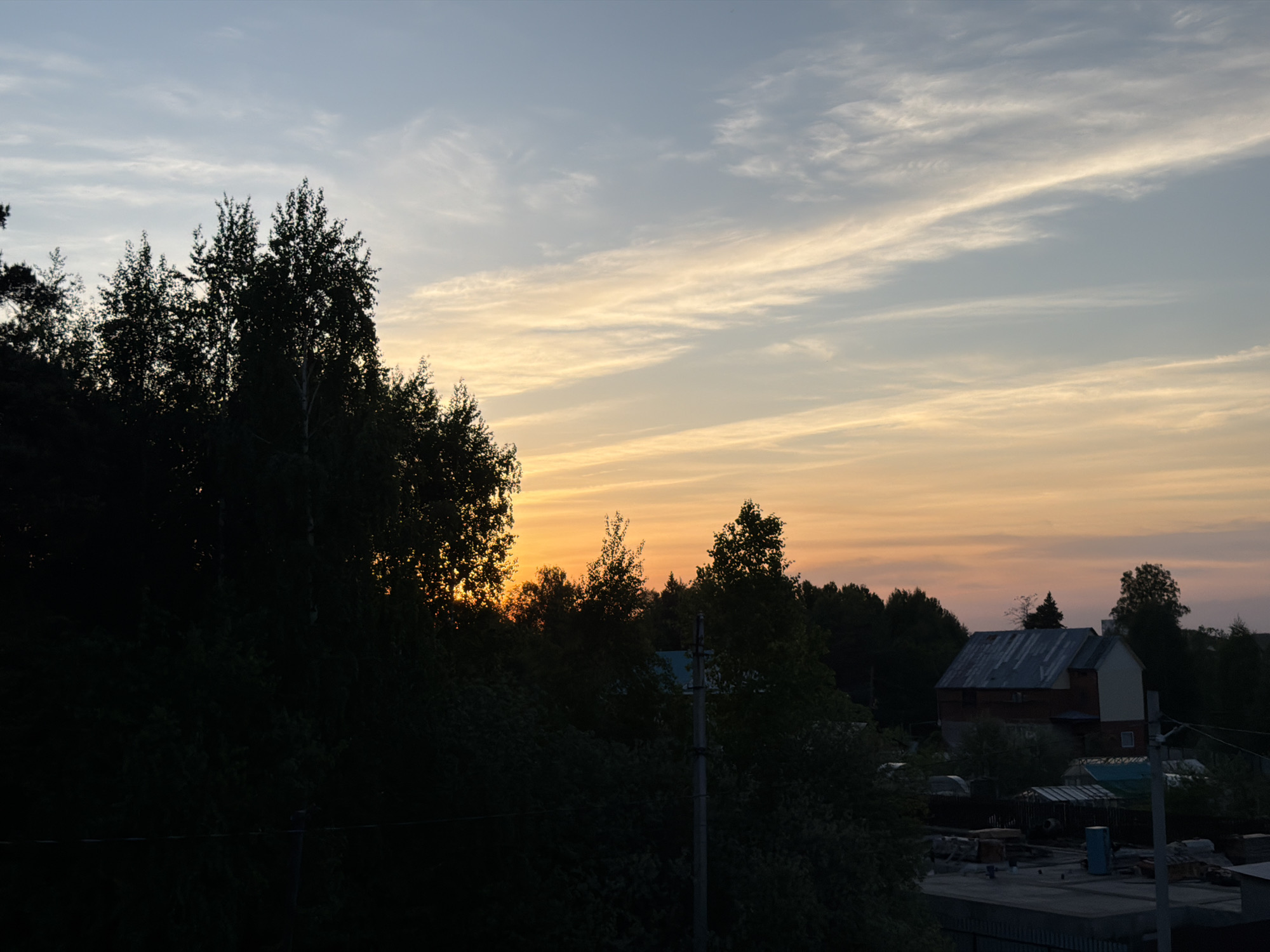 The image shows a sunset view of a residential area in Chaikovsky, Russia. The sky transitions from a light blue to a gradient of yellow, orange, and pink as the sun sets. Thin, wispy clouds are present. Silhouetted trees are in the foreground, with a house visible amongst them. The house has a metal roof and two stories. Fencing and various outdoor items are visible in the lower portion of the image. A power pole is visible in front of the trees.
