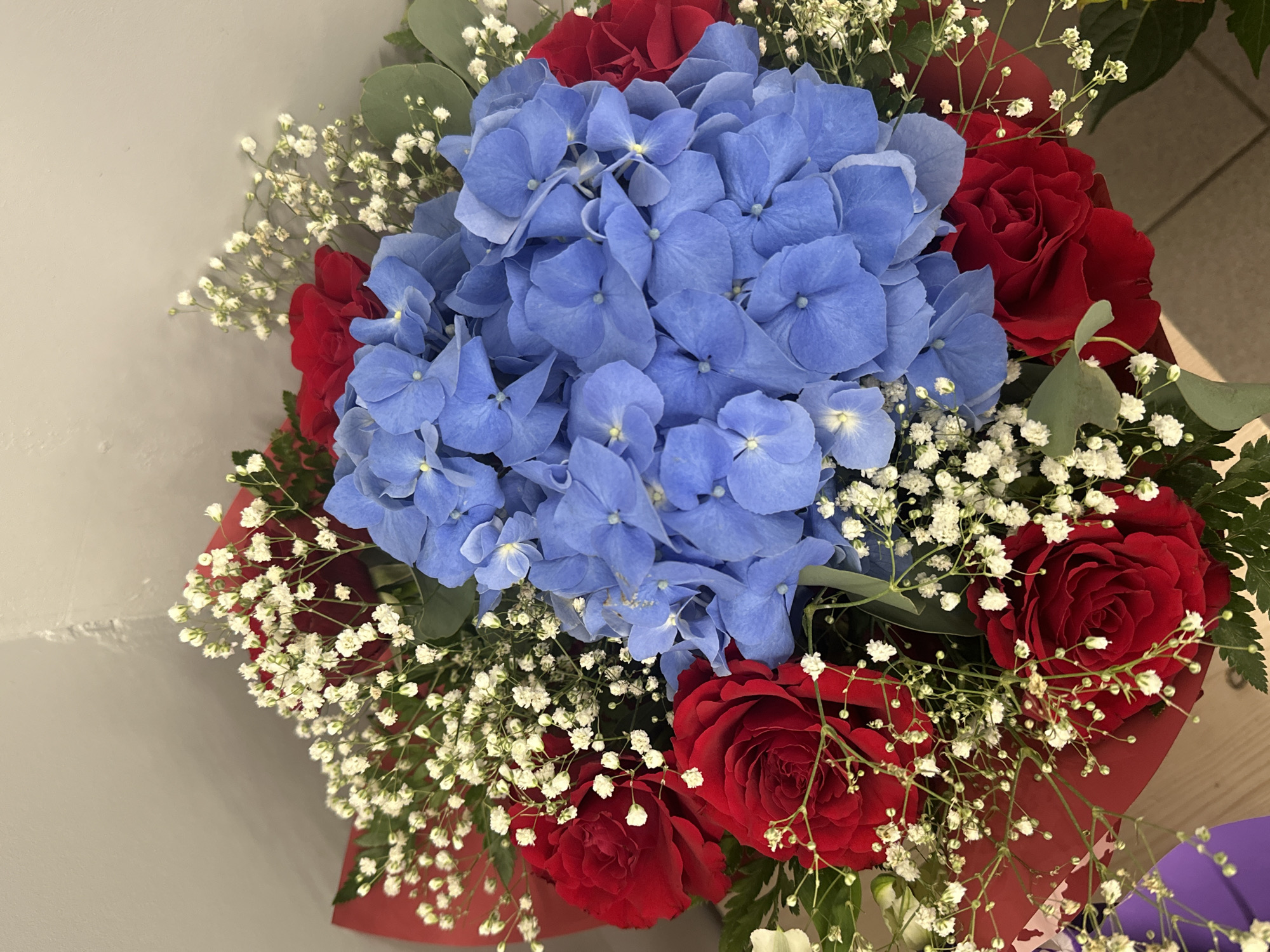 The image depicts a close-up view of a bouquet of flowers. The bouquet features a large blue hydrangea in the center, surrounded by red roses and baby's breath. Green eucalyptus leaves are also interspersed throughout the arrangement. The bouquet is wrapped in red paper. The exact location within Riga, Latvia is not determinable from the provided image.