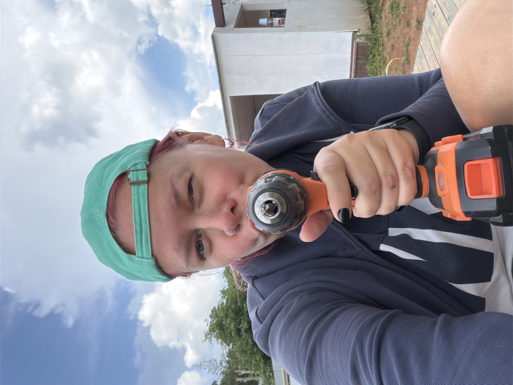 In this image, a person is holding an orange and black drill tool, pointing it towards the camera. The person is wearing a dark blue zip-up jacket over a white t-shirt with black stripes, and a teal baseball cap. Their hair is visible, with pink streaks. They have a nose ring and an earring. The background shows a cloudy sky, trees, and a white building under construction. This scene is likely located in Chaikovsky, Russia, given the context. The overall impression is a candid shot of someone engaged in an outdoor activity.