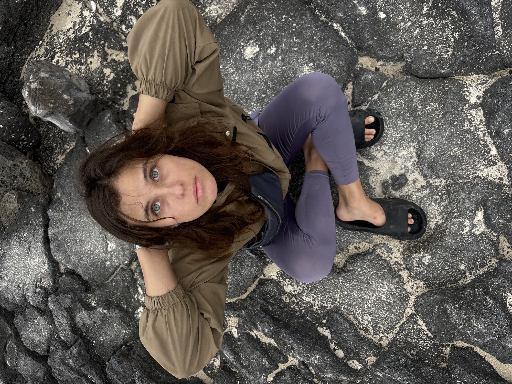 A young woman with long, dark, wavy hair is pictured from a top-down perspective, sitting on dark, textured rocks speckled with white. She is looking directly at the camera with striking blue-green eyes and a neutral expression. She is wearing a light brown jacket with cinched sleeves over a dark blue garment, and purple leggings. Her feet are clad in black slides.
The setting appears to be a rocky coastline, with the dark, volcanic-like rocks and the texture of the sand or coarse ground suggesting a natural, possibly coastal environment. The lighting is somewhat diffused, implying it could be overcast or late in the day, though no strong shadows are visible. The woman's pose, with one arm resting near her head, suggests a relaxed or contemplative mood. The overall composition, with the subject centered against the stark, textured background, emphasizes her presence and direct gaze. There is no visible text in the image. The scene is likely taking place in Jeju, South Korea, given the distinctive rocky terrain often found there.