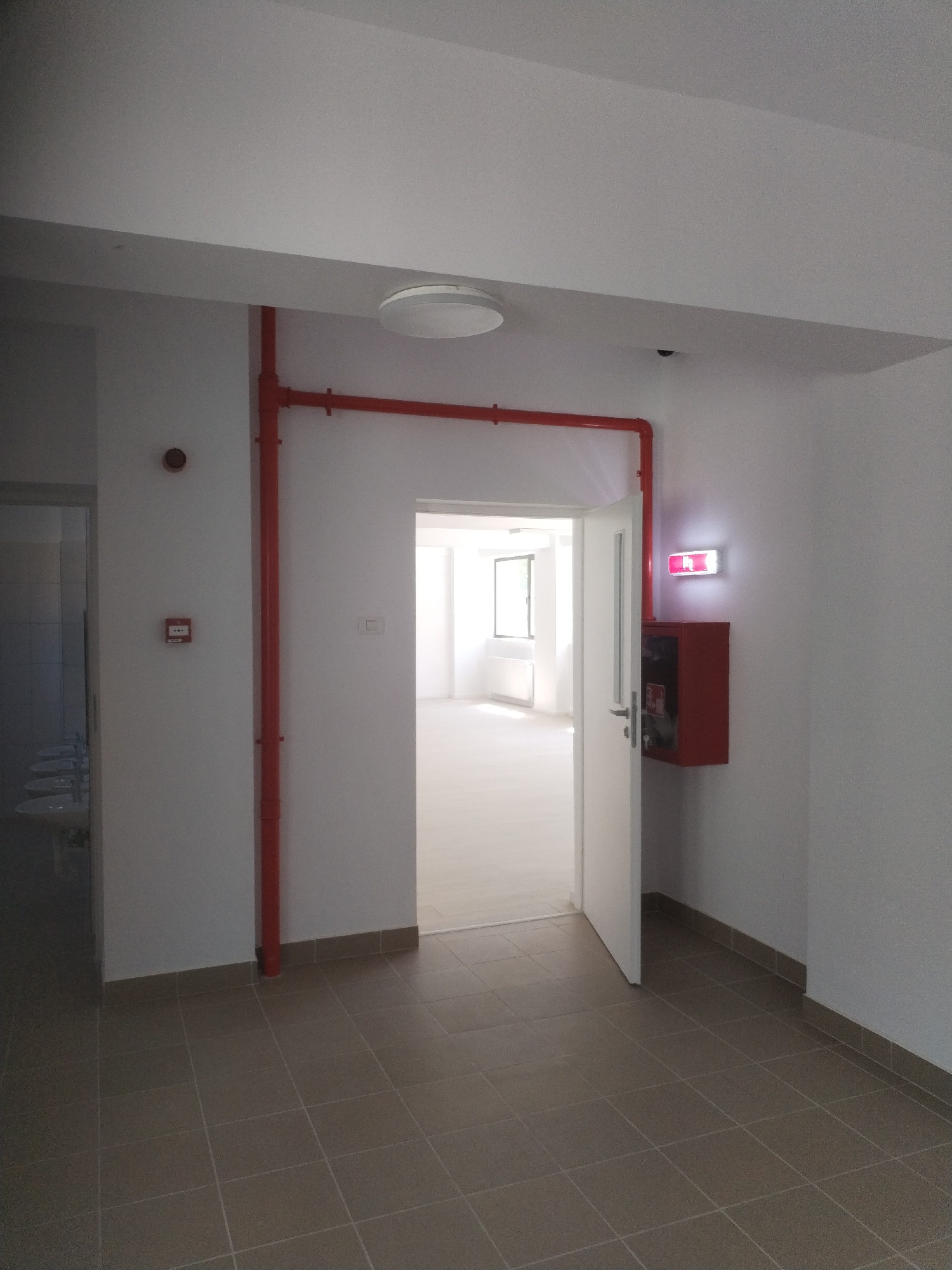 The image depicts an interior view of a building hallway with an open doorway leading into a well-lit room. The hallway features white walls and a tiled floor. Prominently displayed on the wall is a red fire pipe system running vertically and horizontally. To the left of the doorway, a red circular object, possibly a smoke detector, is mounted on the wall, below which is a small red fire alarm box. To the right of the doorway, a red fire extinguisher cabinet is mounted on the wall, with a glowing red emergency exit sign above it. The doorway itself is white with a glass pane, and the open door reveals a bright room with windows and radiators, suggesting a room such as an office or classroom. The environment appears to be a public or commercial building, possibly a school, office, or residential complex. The image is taken from a ground-level perspective.