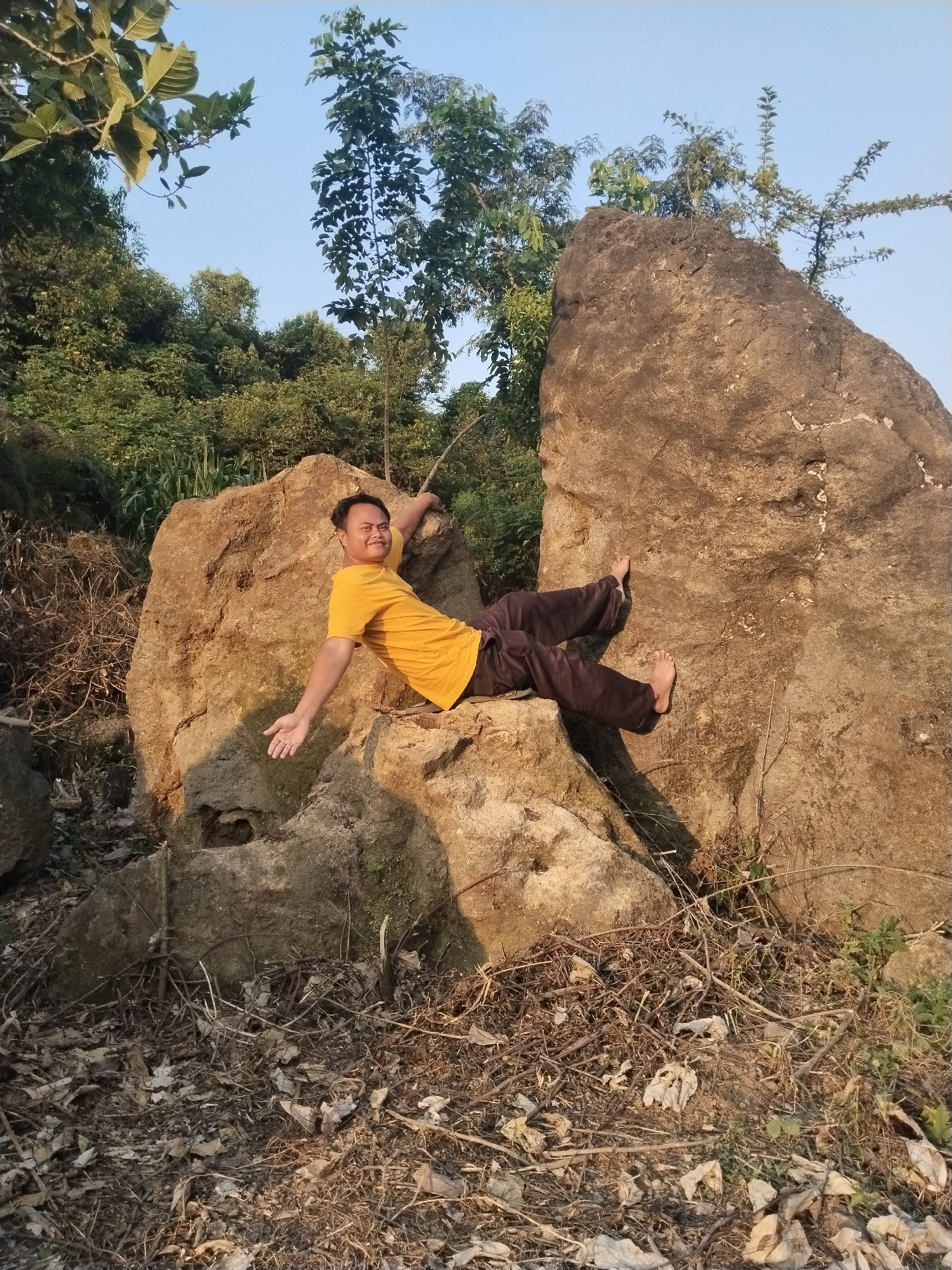 The image captures a smiling man posing on large natural rocks in an outdoor setting in Jambeyan, Indonesia. The subject, an adult male of apparent East Asian ethnicity, is dressed in a vibrant yellow short-sleeved t-shirt and dark pants, and is barefoot. He is leaning against a prominent, light brown boulder, with his right arm bent and resting on the rock while his left arm is extended towards the viewer with an open palm. His legs are bent, with his bare feet propped against another section of the rock. He looks directly at the camera with a broad, cheerful smile, conveying a relaxed and happy demeanor.
The scene is characterized by several massive, rugged boulders that dominate the foreground and midground. The ground surrounding these rocks is covered with a substantial layer of dry leaves, twigs, and fallen branches, suggesting a natural, wild, or unmanaged environment. In the background, dense green foliage, composed of various trees and bushes, indicates a verdant, vegetated landscape under a clear, light blue sky. The lighting suggests either late afternoon or early morning, with warm, golden sunlight illuminating the man and parts of the rocks, casting distinct shadows and creating a soft glow across the scene. There is no visible text within the image.