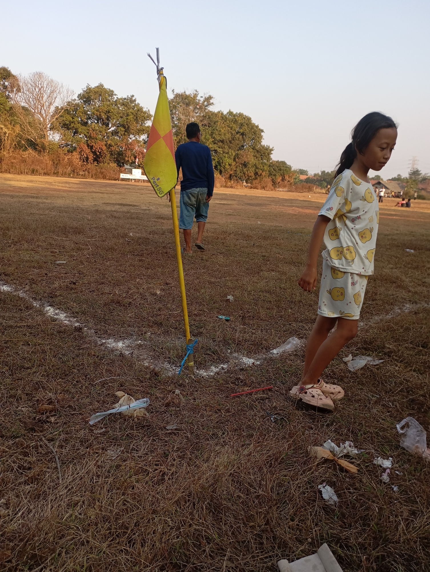 A young girl in a yellow shirt and shorts with a cartoon duck pattern stands on dry, brown grass in an outdoor field. She is looking down and to her left. In the foreground, a yellow flag on a pole stands upright, tied with a blue rope. White lines, possibly marking a field, are visible on the ground. A man in a blue long-sleeved shirt and denim shorts walks away from the camera in the background. Several pieces of trash, including plastic bags and paper, are scattered on the ground. In the distance, trees and some buildings are visible. The sky is clear and pale. This scene appears to be a sports field in Kecamatan Haurgeulis, Indonesia, potentially at dusk.