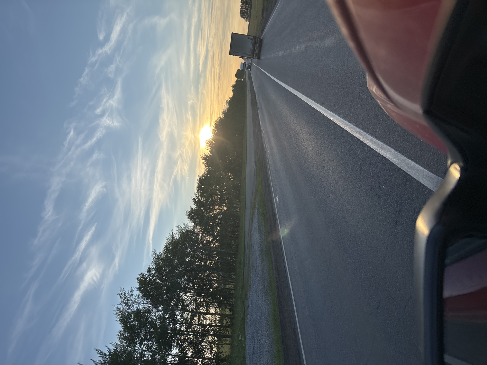 This is a photograph taken from a vehicle, likely a car, looking out at a highway at sunset. The perspective is from the passenger side, with the side mirror and a portion of the car's body visible in the foreground on the right. The road stretches into the distance, marked by a white line on the right lane. Several trucks are visible on the highway, with one in the middle distance and others further ahead.
To the left of the road, a dense line of trees borders the highway, with the setting sun peeking through them, casting a warm, golden light. The sky above is a vibrant blue, streaked with wispy white clouds that are illuminated by the setting sun. The overall impression is one of a peaceful journey during the golden hour, with the natural beauty of the sky and trees contrasting with the man-made road and vehicles. The image evokes a sense of travel and contemplation. The location cues suggest that this scene is taking place in Таймурзино, Russia.