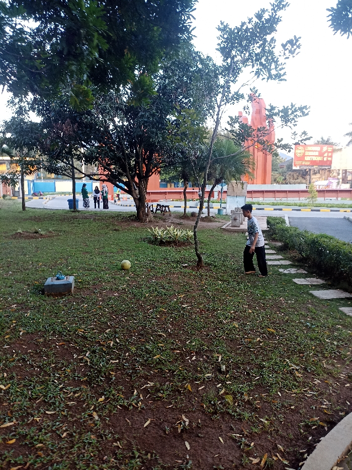 The image shows a grassy area with a large tree in the center and a young man in a blue shirt and black pants standing in the foreground. He is facing the camera, but his back is towards the viewer. There are several people in the distance, dressed in green and blue, walking towards a large, red building in the background. There are dry leaves scattered on the ground. The coordinates provided are the location of the Institut Pertanian Bogor (IPB) campus in Jatinangor, Indonesia. The image shows a grassy area on the campus grounds. The scene is a calm and peaceful one. The man in the foreground appears to be looking at the people in the distance.