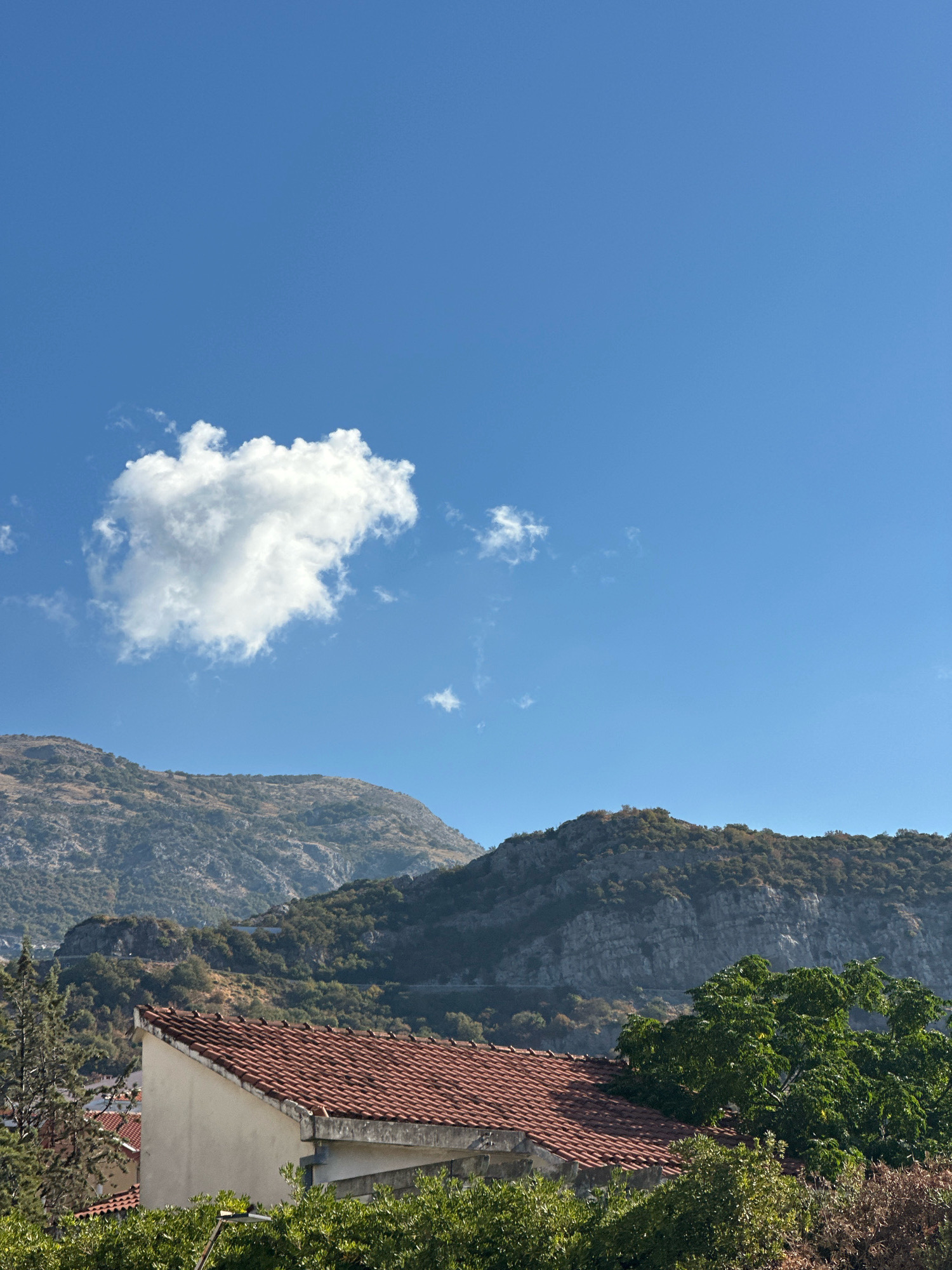 A clear blue sky dominates the upper portion of the image, with a single, puffy white cloud positioned in the upper left quadrant. Below the sky, a mountainous landscape stretches across the frame. The mountains are covered in a mix of sparse vegetation and rocky outcrops, with visible textures and layers. A winding road can be seen traversing the mountainside in the mid-ground. In the foreground, the terracotta tiled roof of a building is prominent. The building appears to be a house or dwelling, with a cream-colored wall visible beneath the roofline. Lush green trees and foliage surround the building, partially obscuring it from view. The scene suggests a sunny day, likely in the late morning or early afternoon, given the bright light and clear skies. The natural beauty of the mountains combined with the presence of human habitation creates a peaceful, scenic view. The image does not contain any visible text. The location context provided suggests this scene is in Budva, Montenegro.