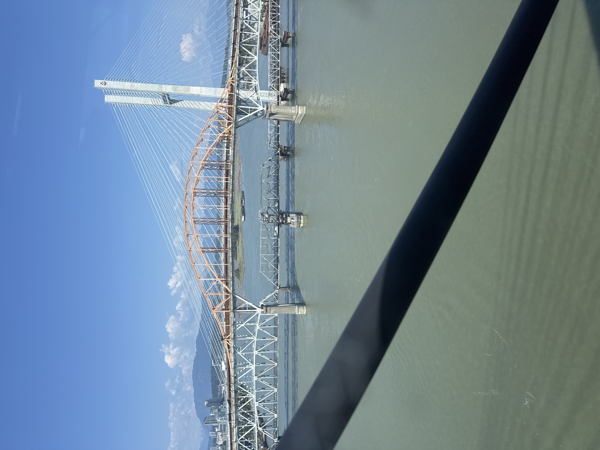 The image shows a view of the Fraser River in New Westminster, Canada, with multiple bridges spanning across it. In the foreground, a dark, diagonal object, possibly a railing or a part of the viewing structure, partially obscures the scene. Beyond this, the river's surface is a murky green, with some visible ripples and disturbances.
Three distinct bridges are visible. The closest bridge is a white, lattice-girder structure supported by concrete piers. To its left, a portion of an orange, arched truss bridge is visible. Behind these, a modern cable-stayed bridge with two tall concrete towers is prominent. This cable-stayed bridge features numerous white cables extending from the towers to the deck. In the distance, a city skyline with buildings and mountains under a clear blue sky with scattered white clouds is visible. Two barges are also present on the river. The overall scene depicts a busy river crossing with a mix of older and newer bridge infrastructure.