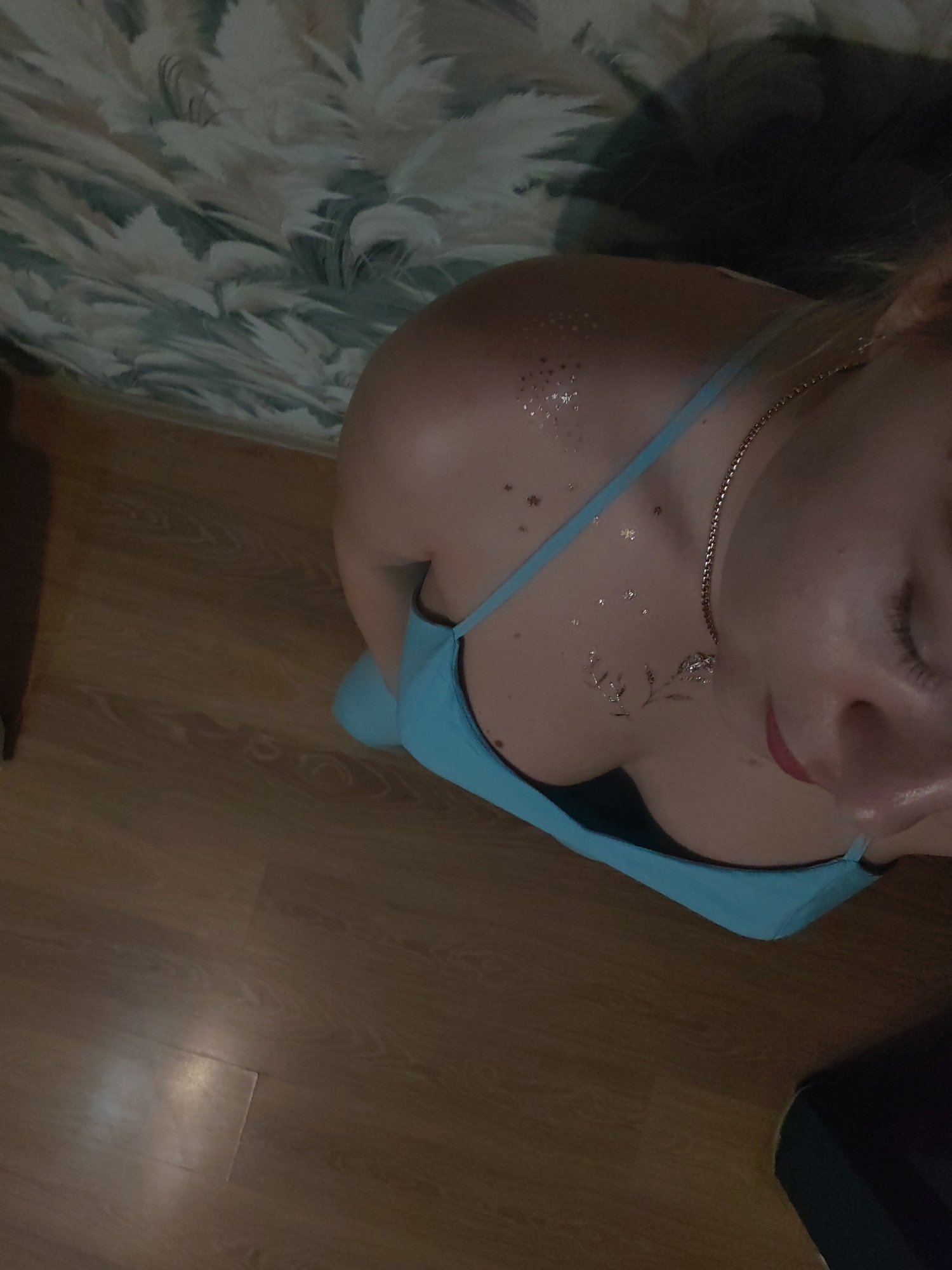 A close-up, high-angle shot shows a person lying down, with their head angled away from the camera. They are wearing a blue dress with thin straps. Glitter or body paint is applied to their shoulder and chest, in the shape of stars and a small branch. A silver chain necklace with a pendant is visible around their neck. The background features a wooden floor and a patterned fabric, possibly a bedspread or curtain, with a light-colored, feathery design. The lighting suggests an indoor setting, possibly in the evening or with dim natural light. The person's face is partially obscured, but their closed eyes and relaxed posture suggest a calm or possibly sleepy state. The setting appears to be a bedroom or living area. The location context is Izhevsk, Russia. There is no visible text in the image.