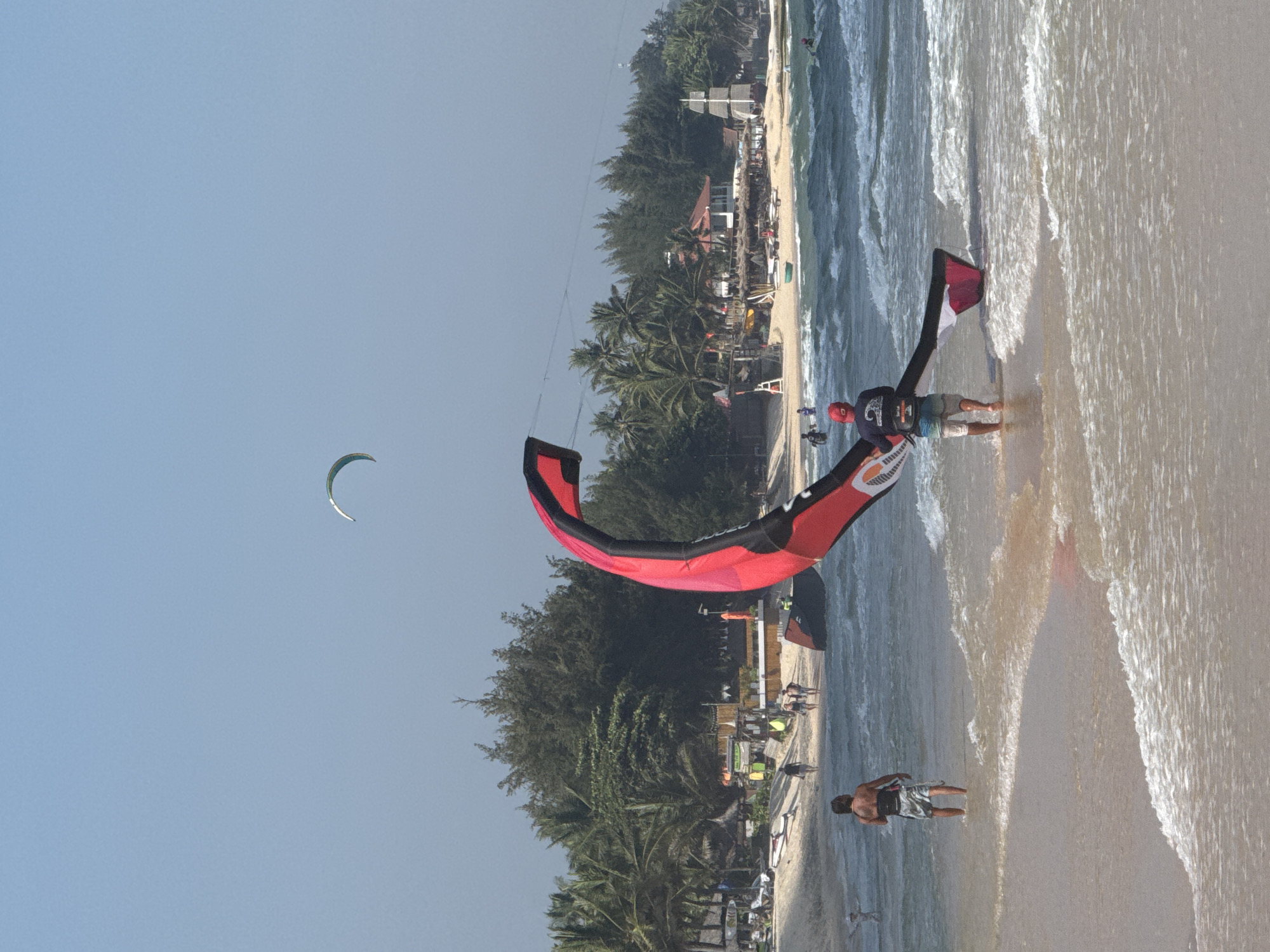 The image depicts a bright, sunny day at a beach in Mũi Né, Vietnam. In the foreground, a person wearing a red cap and shorts is walking in the shallow water, holding a large, predominantly red and black kite. The kite is unfurled and extends out of frame to the right. To the left of this person, another individual in patterned shorts stands facing away from the camera, also in the shallow water.
The middle ground reveals a sandy beach lined with palm trees and other vegetation. Several buildings, possibly beachfront establishments, are visible along the shore, some with thatched roofs. People are scattered on the beach, some walking and others seemingly relaxing. A smaller kite is visible in the sky further in the distance. The sea is a deep blue with moderate waves, indicating windy conditions suitable for kite activities. The overall scene is one of a lively beach environment.
