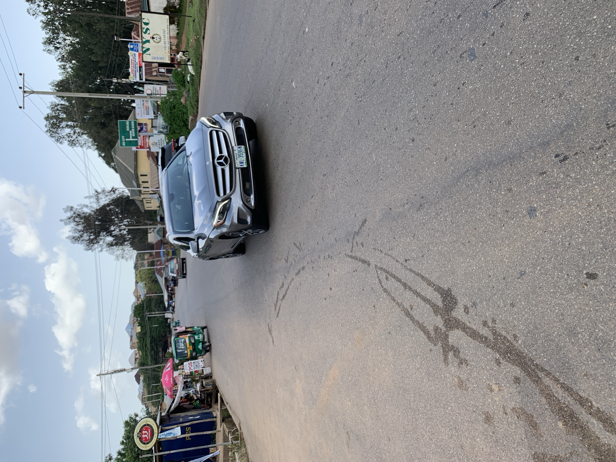 This media file captures a street scene in Jos, Nigeria, under a partly cloudy sky, suggesting it is likely daytime. The foreground is dominated by the asphalt road, marked with tire tracks and what appears to be liquid spills. A silver Mercedes-Benz car is parked on the right side of the road, its license plate reading "KWL-359GN".
Further down the road, a green and yellow tricycle, commonly known as an "Aboki" or "Keke Napep," is visible, indicating local transportation. Along the left side of the road, there are various shops and stalls. Signs visible include "POS" (indicating Point of Sale services), "33 EXPORT," and "NYSC SECRETARIAT" with accompanying text. Another sign in green reads "JOS CITY STAFF SABON BARRI." In the background, buildings and trees line the street, suggesting a moderately developed urban or semi-urban area. The overall impression is a snapshot of everyday life on a Nigerian street.