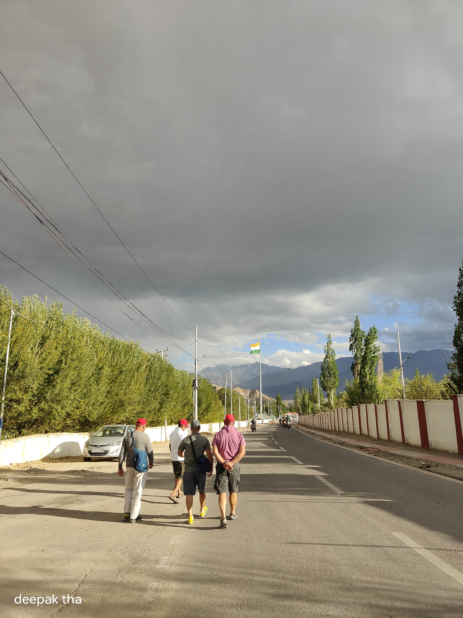 The image shows a group of four men walking away from the camera on a paved road in Leh, India. The sky is overcast with dark clouds, but there are patches of blue sky visible. On the left side of the road, there is a line of green trees and a silver car parked on the side. On the right side, there is a white and red striped wall. Further down the road, a flag is visible on a pole, which appears to be the Indian flag. Mountains can be seen in the background. The men are dressed in casual clothing. One man on the far left is wearing a gray long-sleeved shirt and white pants. The second man from the left is wearing a dark gray t-shirt and shorts, with a blue bag slung over his shoulder. The third man from the left is wearing a pink t-shirt and shorts, and the fourth man, on the far right, is wearing a maroon shirt and shorts. All four men are wearing red caps. The scene is likely captured on a road leading into or out of the main city area of Leh, given the presence of the Indian flag and the mountainous landscape.