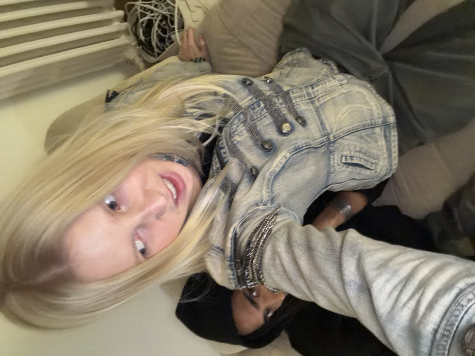 An indoor scene in the city of Paris, France, depicts two individuals in close proximity. In the foreground, a fair-skinned individual with long, straight blonde hair, parted in the middle, faces slightly away from the camera, looking towards the viewer's right with a grimacing expression, mouth slightly open, and eyes wide. This individual wears a light-wash denim jacket with military-style detailing, including horizontal bands of small metallic studs or beads across the chest and decorative chains draped over the left shoulder. A black garment is visible underneath, and a silver-toned cuff or bracelet is on their right wrist. Their left arm extends forward, suggesting they are holding the camera. Behind and to the right, a second individual with darker skin is partially visible, wearing a black hooded garment with the hood pulled up. Only their right eye and a portion of their face are discernible, looking directly towards the camera with a neutral expression. Both appear to be seated on a light-colored, upholstered couch or armchair. The background shows a plain light-colored wall, a section of a white corrugated radiator, and a light-toned bag containing tangled white and dark cables. The lighting is artificial.