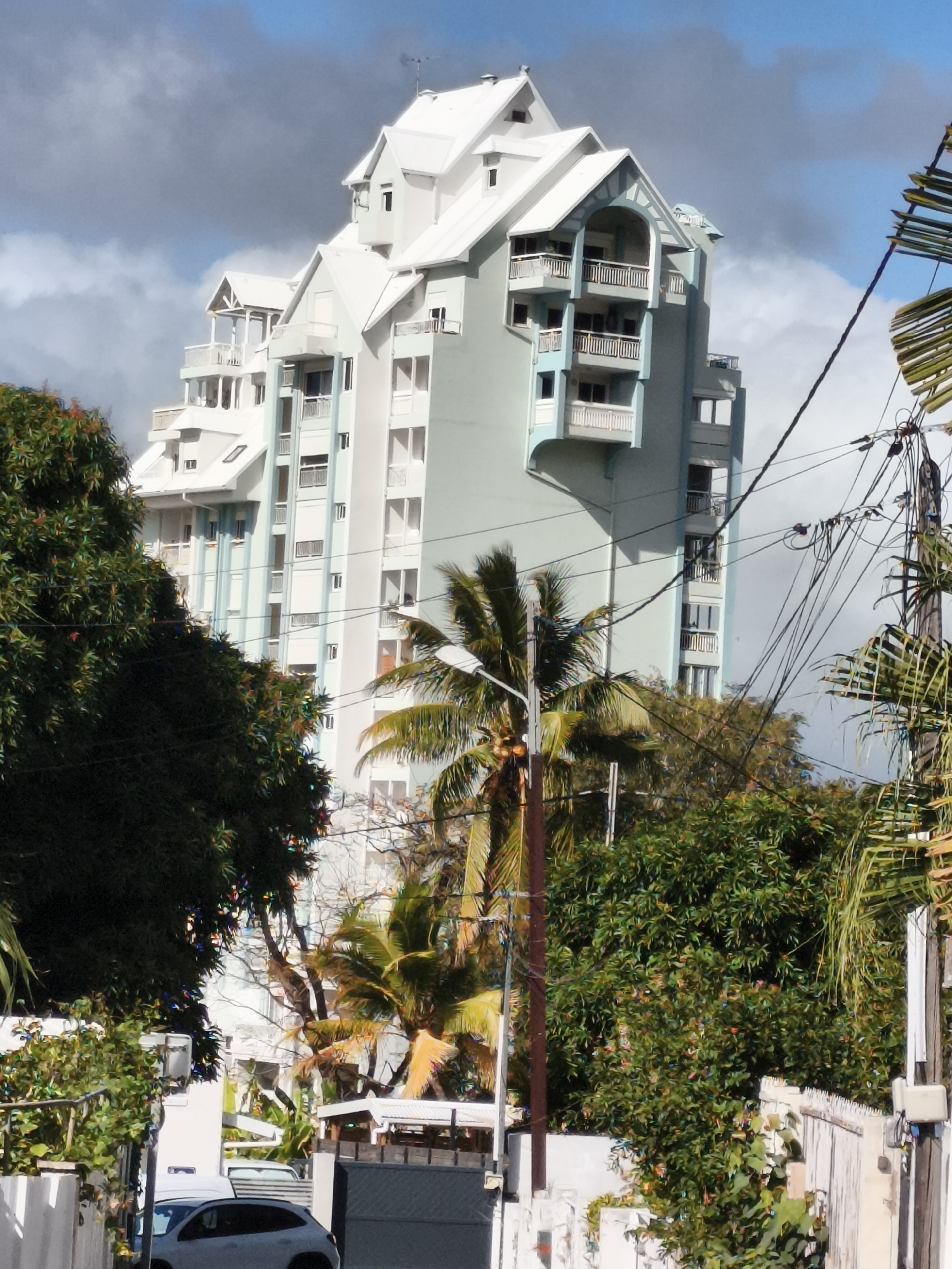 This is a photograph taken from a low angle, looking up at a multi-story building with a distinct, tiered architectural design, likely a residential complex. The building's exterior is primarily light blue and white, with multiple balconies and white railings. The roofline is complex, featuring several gables and slopes, giving it a somewhat whimsical or ornate appearance.
To the left of the building, a large, dark green tree dominates the foreground, its leaves dense and obscuring some of the lower parts of the structure. To the right, a utility pole with a complex network of black wires is visible, along with palm fronds and other lush green foliage. In the lower part of the image, a white fence runs along the right edge, and a white car and a dark fence are visible at the very bottom, suggesting a street or residential area.
The sky is partly cloudy, with patches of blue sky peeking through grey clouds, indicating dynamic weather. The overall impression is one of a tropical or subtropical urban environment, given the presence of palm trees and dense greenery.
The user states the location is Saint-Denis, France. This detail is important as Saint-Denis, France is a city in the Île-de-France region, north of Paris. However, the architectural style, lush vegetation, and palm trees in the image are not typical of mainland France. This imagery is more consistent with Saint-Denis, Réunion Island, a French overseas department located in the Indian Ocean, where such architecture and flora are common. Therefore, assuming the user meant Saint-Denis, Réunion Island, the image depicts a building in that location.