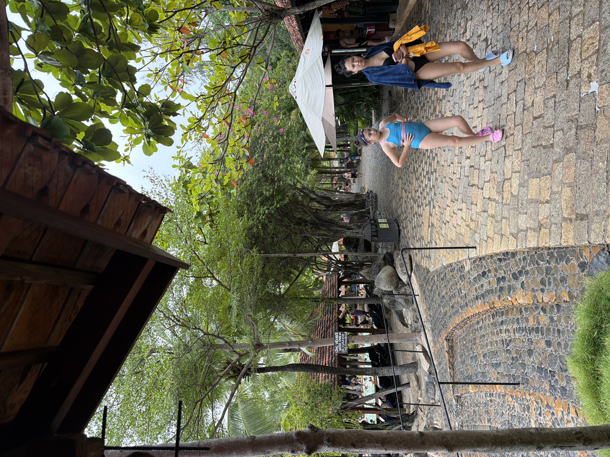 The image depicts a pathway in a tropical resort, likely in Nha Trang, Vietnam, given the presence of lush vegetation and the water park signage. The pathway is paved with cobblestones and flanked by trees and foliage. In the foreground, two females, an adult and a child, are walking on the path. The child is wearing a blue swimsuit and pink sandals, while the adult is wearing a black bikini, a blue towel draped over her shoulder, and holding a yellow towel and a pink smartphone. Behind them, several other people can be seen, some near a swimming pool and others relaxing under thatched-roof structures. The environment suggests a leisure or recreational setting, possibly a water park. The overall atmosphere is casual and summery.