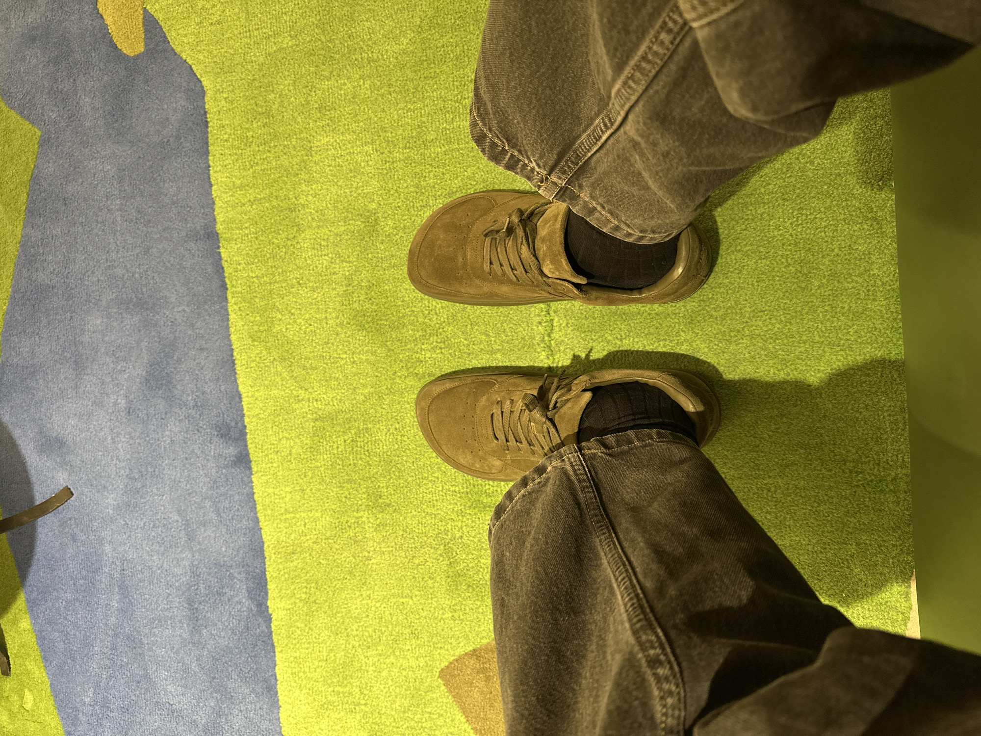 A first-person perspective shot looking down at a person's legs and feet. The person is wearing dark gray jeans and olive-green suede sneakers with black socks. The background consists of a green, blue, and yellow patterned carpet. The floor appears to be level. No other persons are visible in the image. There is no identifying information to suggest that this photo was taken in London, United Kingdom.