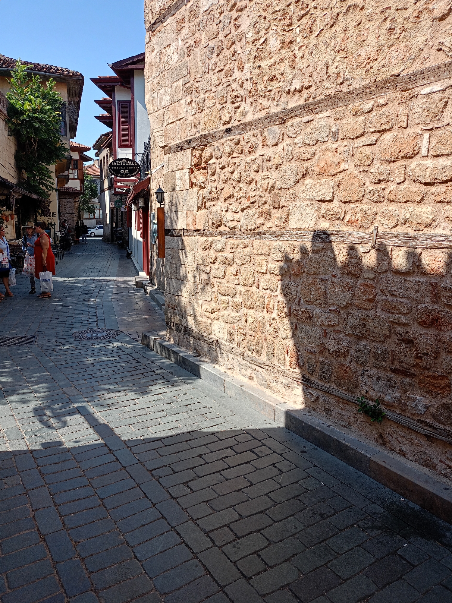 The image depicts a narrow, cobblestone street in Kaleiçi, Antalya, Turkey. The street is lined with old stone buildings, one of which has a rough, textured stone facade. Shadows of people and foliage are cast across the cobblestones. In the background, a white car is parked, and other buildings with tiled roofs are visible. A sign for "SAINT PAUL CULTURAL CENTER" hangs from one of the buildings. Several people are walking on the street, some carrying shopping bags. The lighting suggests it is a sunny day.