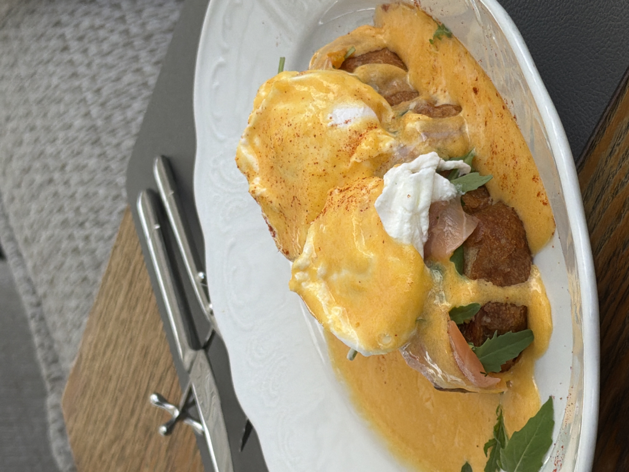 The image shows a close-up, overhead view of a dish of Eggs Benedict served on a white, ornate plate. The dish consists of two poached eggs topped with hollandaise sauce, seasoned with paprika. Beneath the eggs are pieces of toasted bread, topped with what appears to be cured salmon and some greens. The plate is set on a dark gray surface, and a wooden table is visible in the background, with a pair of silver cutlery resting on it. The scene is lit, suggesting it is an indoor setting, possibly a cafe or restaurant. There is no indication of the specific location within Saint Petersburg, Russia, beyond the context provided.
