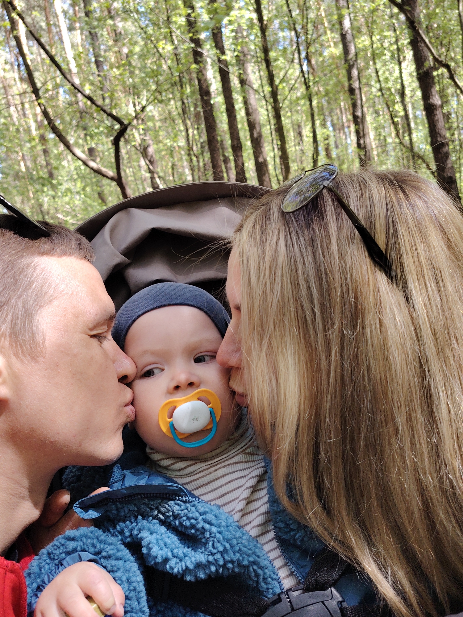 The image is a close-up shot of a young family of three in a forest setting. A baby, wearing a blue hat and jacket, is centrally positioned with a pacifier in its mouth. The baby's parents are on either side, each kissing one of the baby's cheeks. The father is on the left, with short hair, and the mother on the right, with long blonde hair and sunglasses resting on her head. The background is a forest of tall trees with green leaves, suggesting it's a sunny day. The photo appears to be taken from inside a stroller or a carrier. There is no information in the image to place it in the city of Chaikovsky, Russia, or any other location.