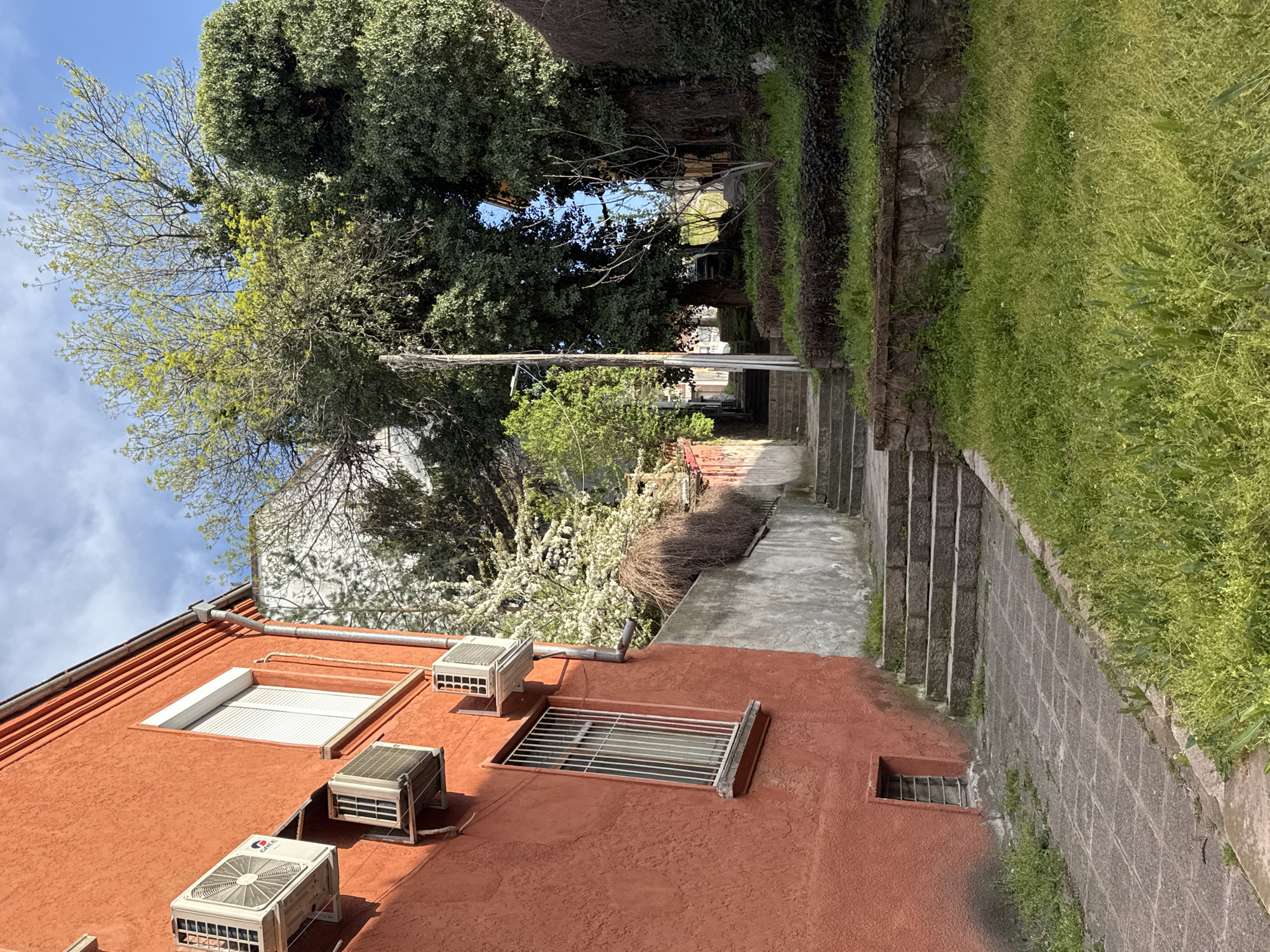 This photo captures an outdoor scene in Plovdiv, Bulgaria, looking up a narrow walkway framed by buildings and greenery. The walkway is paved with stone and ascends via steps along the right side. A red building is visible on the left, featuring windows protected by metal bars and several air conditioning units mounted to its exterior. Above the building, trees with varying degrees of foliage fill the frame. The sky is visible in the top portion, exhibiting a mix of clear blue and scattered clouds. A utility pole is visible among the trees. The lighting suggests it is daytime, with shadows cast by the buildings and foliage. No people are readily discernible in the scene. The environment is a blend of urban and natural elements.