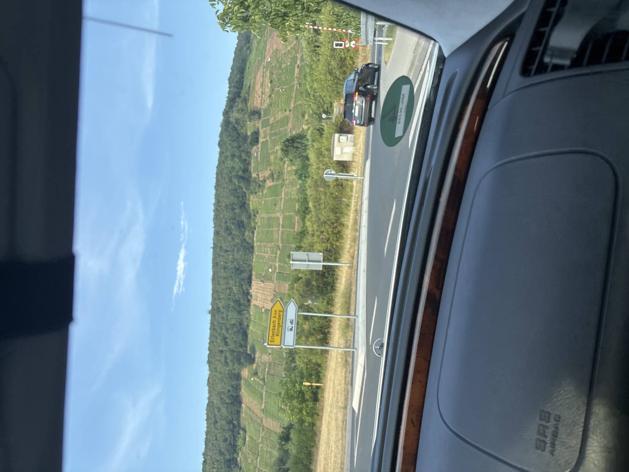 The image shows a view from inside a car, looking out at a rural landscape. In the foreground, the dashboard of the car is visible, with the "SRS AIRBAG" logo clearly imprinted on it. The car appears to be at a road intersection.
In the middle ground, there is a road with white lane markings. To the left, there is a yellow directional sign indicating "Erlenbach 4 km" and "Klingenberg" with arrows pointing to the left and straight ahead, respectively. Next to the directional sign are two other signs: one round sign with a bicycle symbol and a car symbol, and a rectangular sign with a train symbol. Further to the right, a black SUV is visible turning right at the intersection. A red and white striped railway crossing barrier is also present on the right side of the road.
In the background, a hillside covered in vineyards is prominent, with rows of grapevines clearly discernible. The sky is blue with scattered white clouds. The overall environment suggests a sunny day in a wine-growing region.
The location is the city of Wörth am Main, Germany, specifically at an intersection leading towards Erlenbach and Klingenberg.