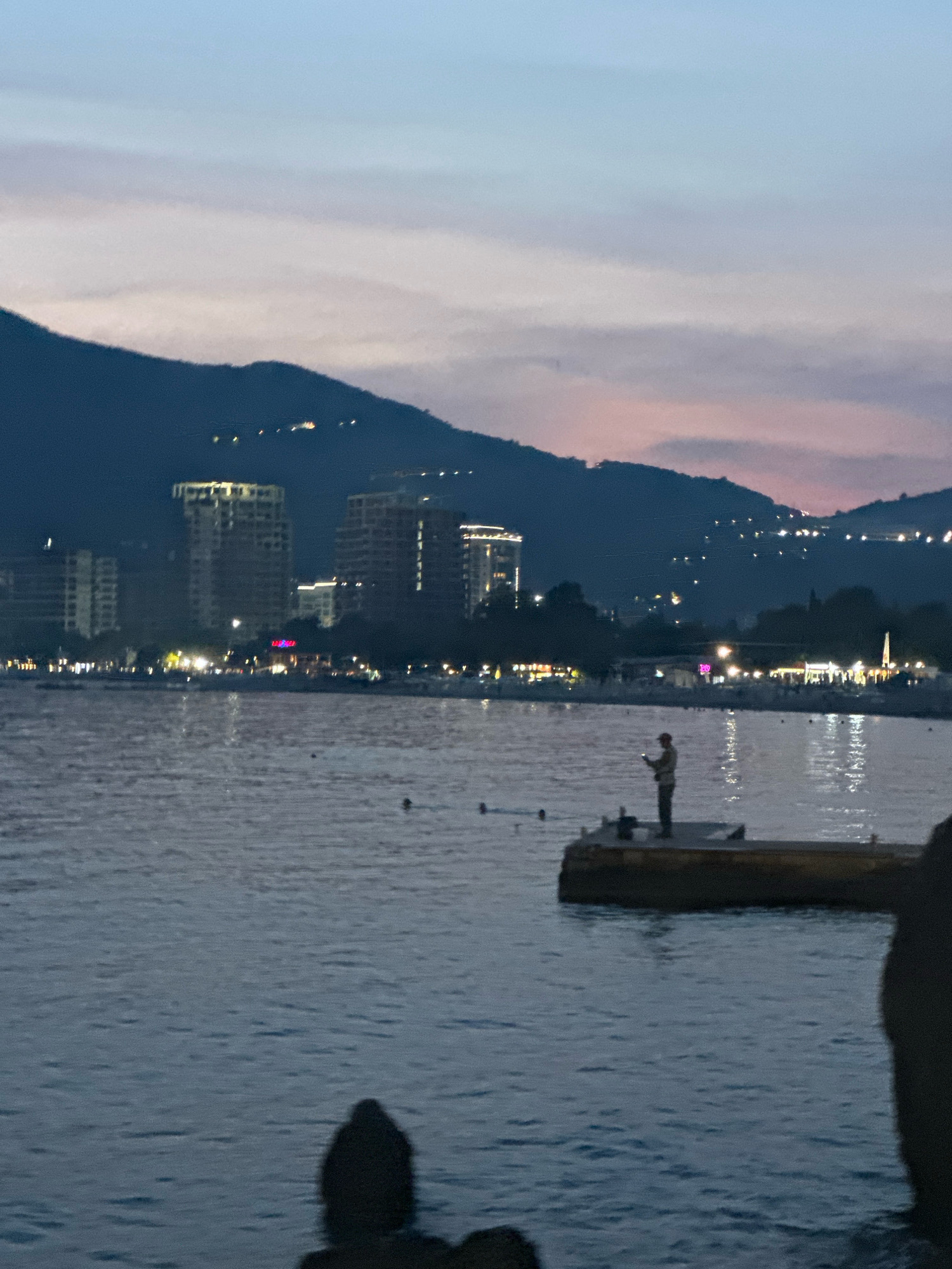 A long shot captures the coastline of Budva, Montenegro, at dusk. The water in the foreground is calm, reflecting the fading light. A single person stands on a small pier, fishing. In the background, the Budva skyline is illuminated by streetlights and building lights, with tall buildings and a mountain range behind them. The sky is a gradient of blues and purples, with hints of pink from the setting sun. A silhouetted figure is visible in the very front of the image.