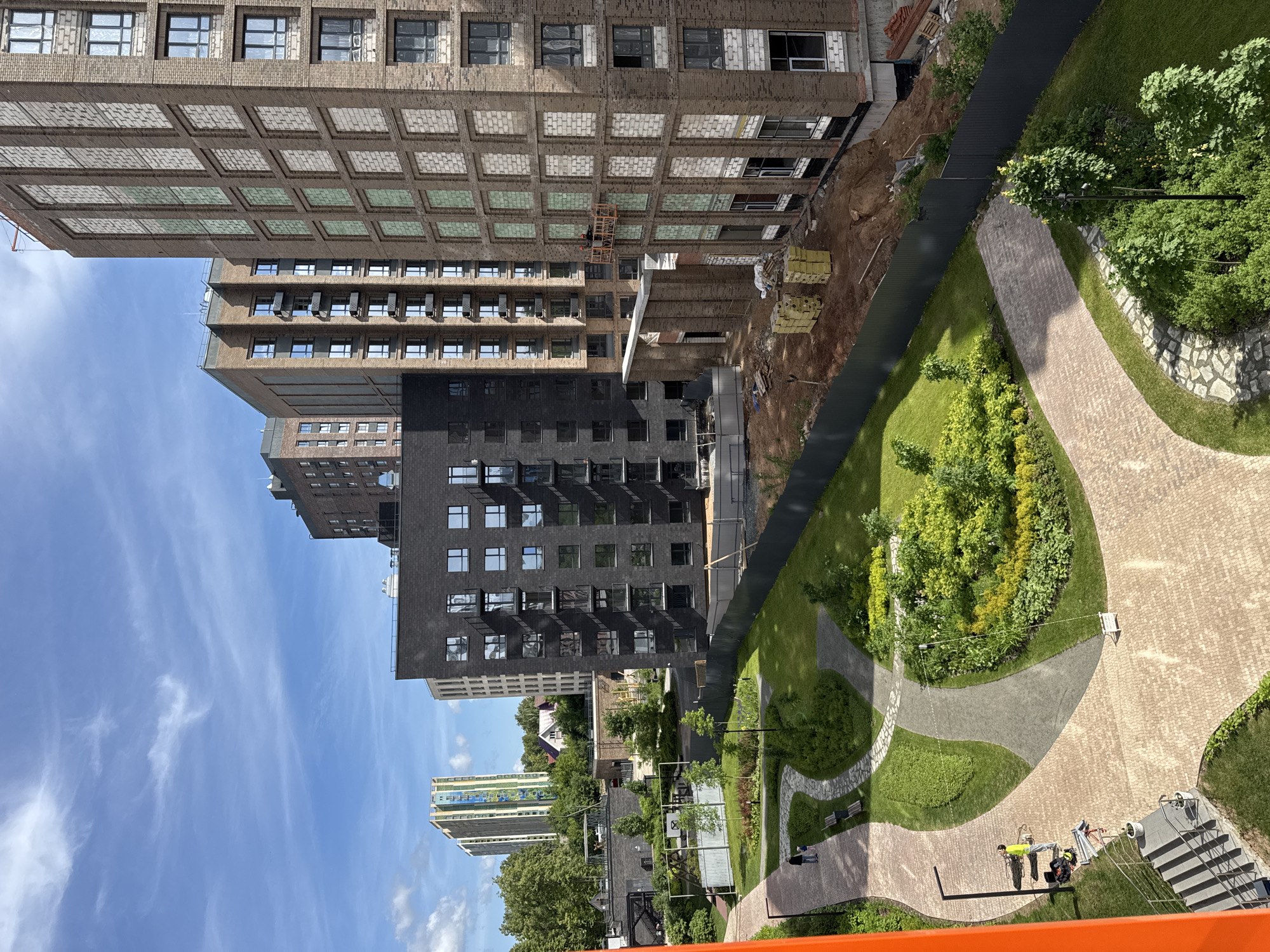 This image presents an aerial view of a modern residential development in Izhevsk, Russia, identifiable by the distinctive multi-colored high-rise building resembling the Kvartalny Residential Complex visible in the distant background on the left.
In the foreground, an orange structural element, possibly part of a building's balcony or frame, partially obscures the lower-left corner. Below this element, a set of outdoor stairs with a metal handrail descends. Adjacent to the stairs, a person in a bright yellow shirt and dark pants stands, attending to equipment including cables and a tripod, positioned on the paved pathway.
The scene is dominated by a newly landscaped park area featuring winding pathways composed of light brown pavers and sections of grey cobblestone/pavers. This area includes well-maintained green lawns, numerous young trees, shrubs, and ornamental plants. Several dark-colored benches are placed along the paths. In the mid-left background of the park, a basketball hoop and court are visible behind a light grey fence, and further back, a small playground with yellow structures is discernible. Another person and a child walk along a paved path in the upper-left section of the park.
To the right of the park area, an active construction zone is delineated by a dark grey metal fence. This zone comprises exposed reddish-brown earth with piles of dirt and scattered construction debris. Stacks of yellow-wrapped building materials are visible on the ground.
Several multi-story residential buildings populate the scene. On the far right, a tall, multi-story building with a brown/orange brick facade is undergoing construction. Many of its window openings are filled with white/light grey blocks, and some completed windows are covered with protective film. Two construction workers, wearing orange vests and hard hats, are visible on an orange suspended scaffolding platform, positioned midway up the building's facade. Behind this building, another brick building of similar design is partially visible. Adjacent to the construction area and behind the park, a modern, dark grey/black building stands, featuring numerous large windows and dark-colored balconies, appearing largely complete. Further back and to the left of the dark building, additional residential structures with lighter facades are visible.
The sky is clear blue with scattered wispy white clouds. The overall impression is one of ongoing urban development, blending new construction with recently completed residential units and green spaces.