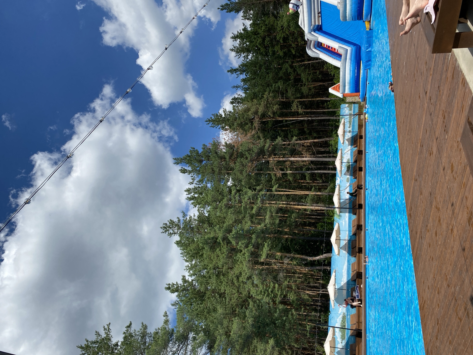 The image depicts an outdoor swimming pool area at a resort, likely in the city of Voronezh, Russia. The pool is a large, rectangular body of bright blue water. Wooden decking surrounds the pool, with lounge chairs and umbrellas set up along one side. In the background, a dense forest of tall pine trees creates a natural backdrop. A large inflatable water slide, predominantly blue and white, is situated at the far end of the pool. Several people are visible in and around the pool, some relaxing on lounge chairs, others swimming, and at least one person appears to be on the water slide. String lights are hung across the upper portion of the image, suggesting the area is also used in the evening. The sky is partly cloudy with a clear blue sky visible in sections.