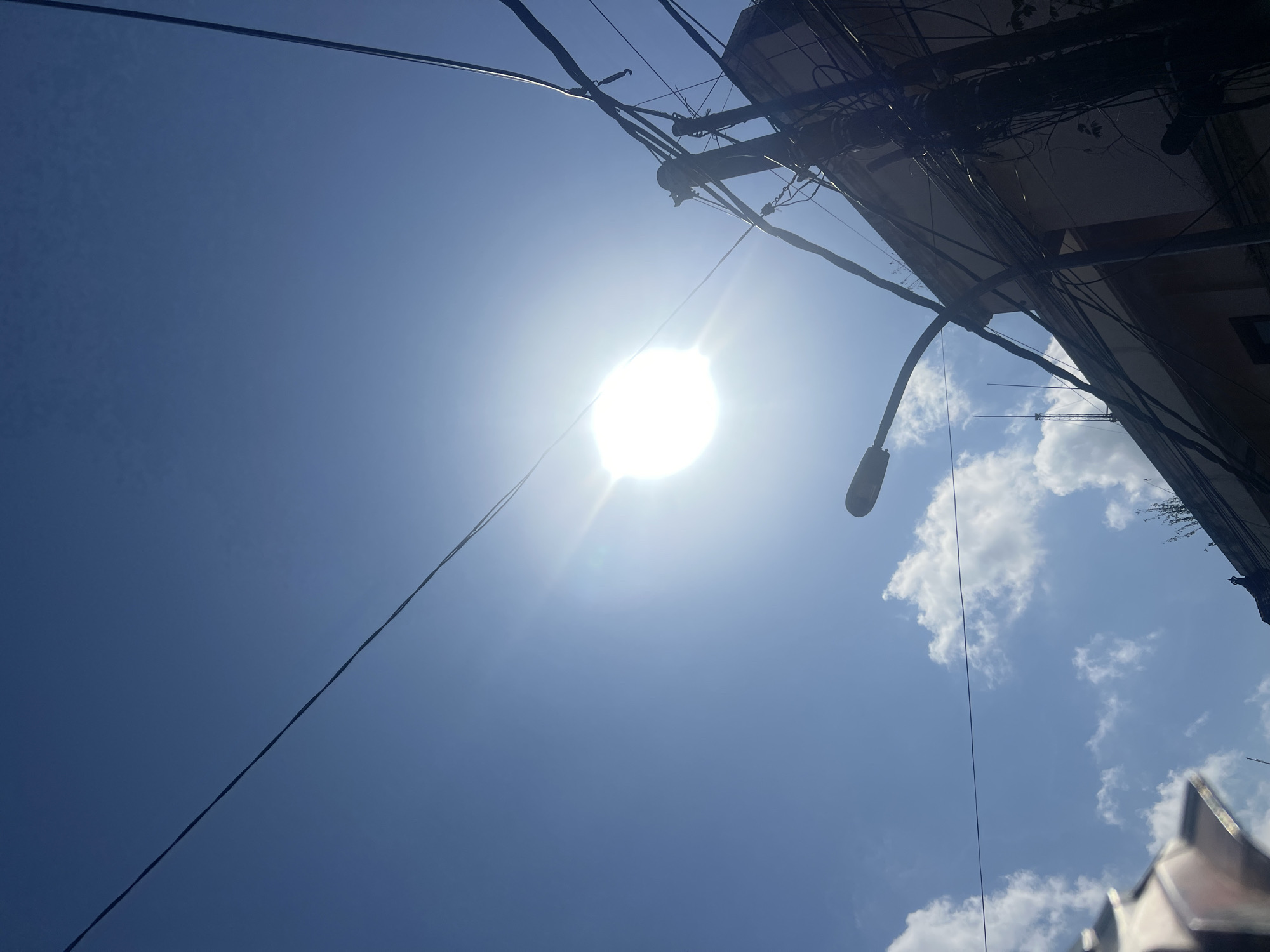 An upward-looking, wide-angle shot captures a bright, sunlit sky over Kuta, Indonesia. The sun is a brilliant, blown-out orb in the upper center of the frame, casting strong lens flare and washing out the surrounding blue sky. A power pole with a tangle of black electrical wires crisscrosses the upper right portion of the image, obscuring parts of a light-colored building peeking from the bottom right corner. A streetlamp arm extends from the left edge of the frame towards the sun. Scattered white clouds drift across the sky. The scene suggests a typical urban environment with overhead power infrastructure.