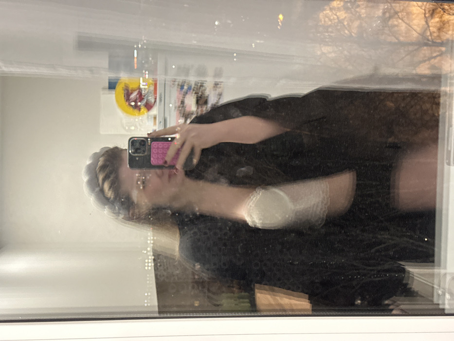 A person holds a cell phone with a pink, bubble-textured case, taking a selfie in a mirror. The reflection is somewhat blurred. The person is wearing a dark, textured top or dress and has a headband. In the background, there are pictures and various items on a wall. The scene is illuminated by daylight coming through a window, reflecting the outside.