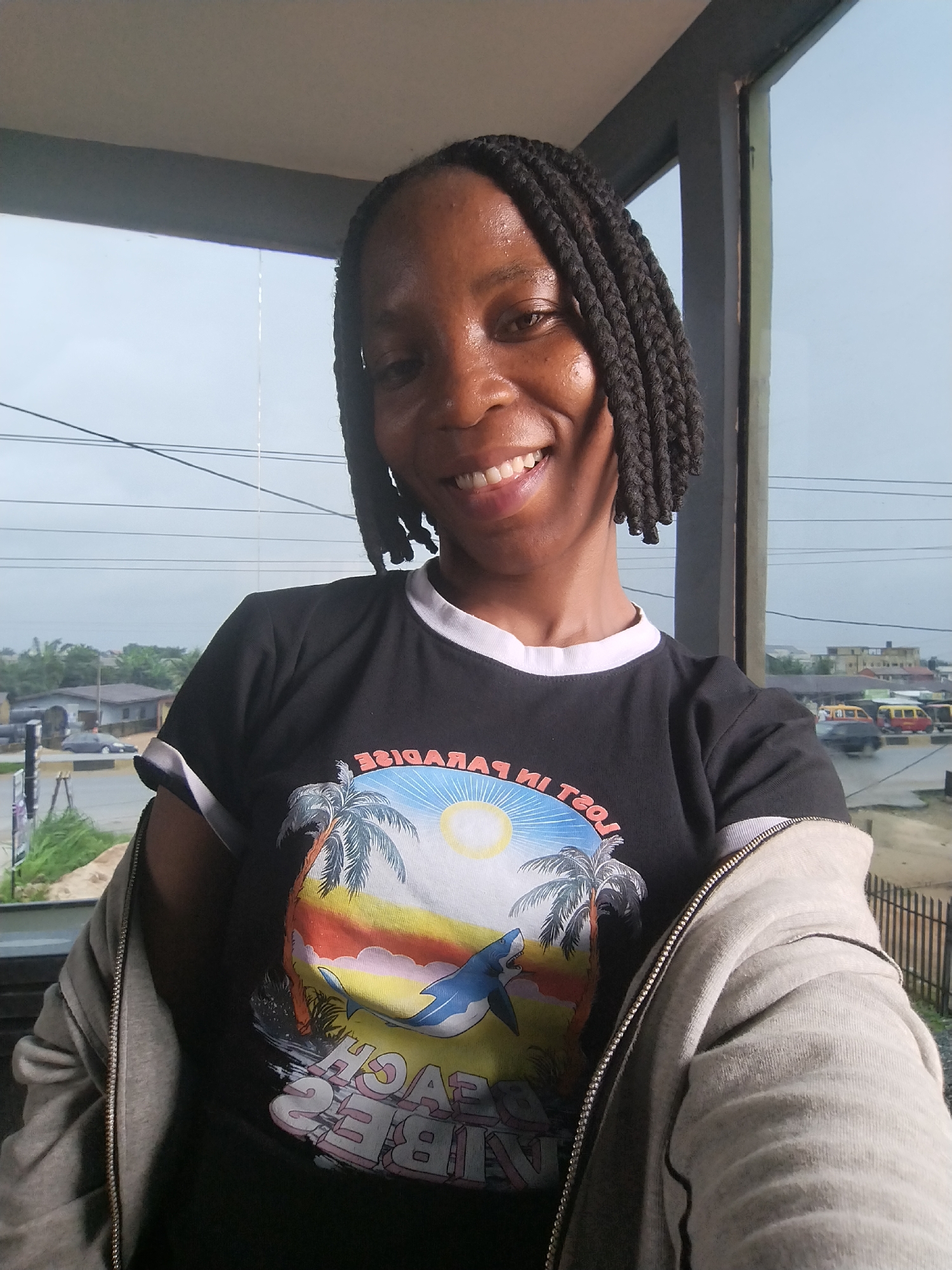 A medium shot captures a Black woman with dark, braided hair, smiling at the camera. She is wearing a black t-shirt with a graphic print of a tropical scene featuring palm trees, a sun, and a shark. She also wears a light gray zip-up jacket over her t-shirt. The background appears to be an outdoor setting, possibly a veranda or balcony, with a view of a street with buildings, vehicles, and vegetation. The image is taken with a slight upward angle, and the lighting suggests an overcast day. The overall impression is informal and candid. While the exact location within Uselu, Nigeria cannot be precisely determined from the visual information alone, the scene depicts an urban environment consistent with many Nigerian cities.