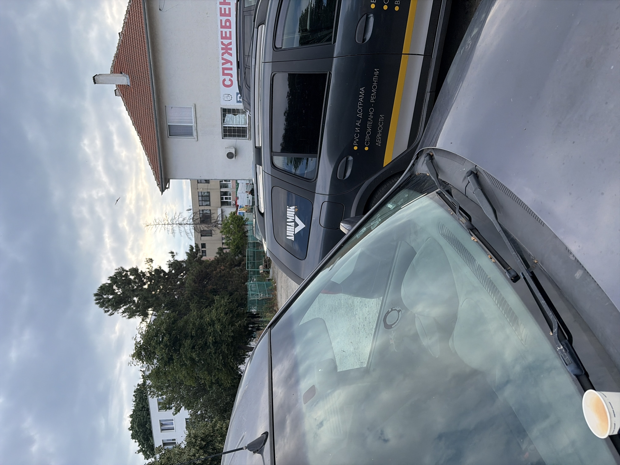 The image captures a cloudy sky over a street scene in Burgas, Bulgaria. In the foreground, the hood and windshield of a dark grey car are visible, with a white paper cup resting on the lower left of the windshield. Reflected in the windshield are clouds and the sky. Behind this car, another dark grey station wagon is parked, bearing logos and text in what appears to be Cyrillic script. One logo reads "ДИНАМИК" (Dinamik), and another section of text states "• PVC И AL ДОГРАМА" (PVC and AL joinery) and "• СТРОИТЕЛНО - РЕМОНТНИ ДЕЙНОСТИ" (Construction - repair activities). A white building with a red-tiled roof and the partial word "СЛУЖЕБЕН" (Official) is visible on the right side of the frame. Further in the background, trees and other buildings can be seen. The overall atmosphere is overcast, suggesting potentially inclement weather.