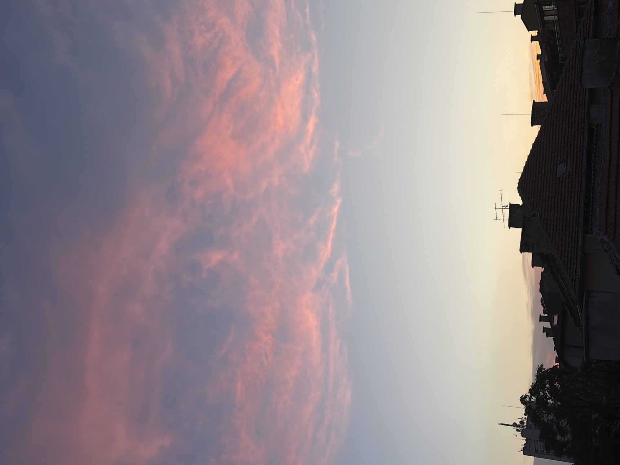 The image shows a cityscape at dusk, with a wide expanse of sky dominating the upper portion. The sky is filled with wispy clouds tinged with shades of pink and purple, transitioning to a pale blue at the top. Below the sky, silhouetted against the fading light, are the rooftops of buildings. Some buildings appear to be residential, with tiled roofs and chimneys. A taller building with antennas is visible on the left side of the frame, adding a modern element to the urban landscape. The overall atmosphere is serene and tranquil, characteristic of twilight. The location is identified as Unknown, Unknown.