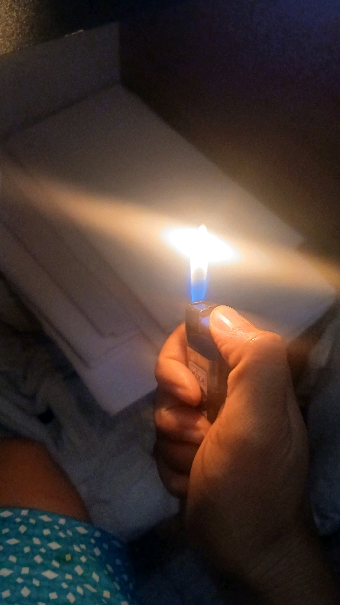 A person's hand holds a lit lighter, producing a bright flame. The background appears to be indoors with some indistinct objects that could be furniture or boxes. There is a blue and white patterned fabric visible in the lower left of the image. The lighting suggests it is nighttime or a dimly lit room. The location of Gura Top, Nigeria is not determinable from the image.