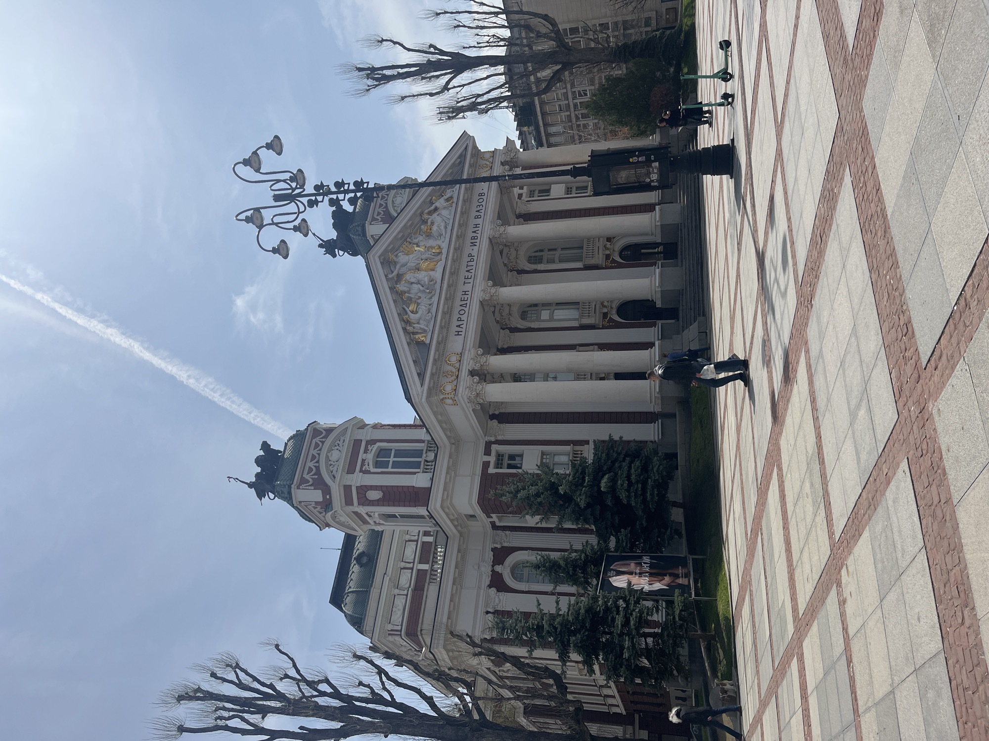 The image shows the Ivan Vazov National Theatre in Sofia, Bulgaria. The building is visible with its classical facade, including columns and statues. A lamppost with multiple lights stands in front of the building. There is a square in front of the theatre, paved with square tiles with red accents. A person is walking along the pavement. Bare trees are visible in the background. The sky is pale blue with a contrail.
