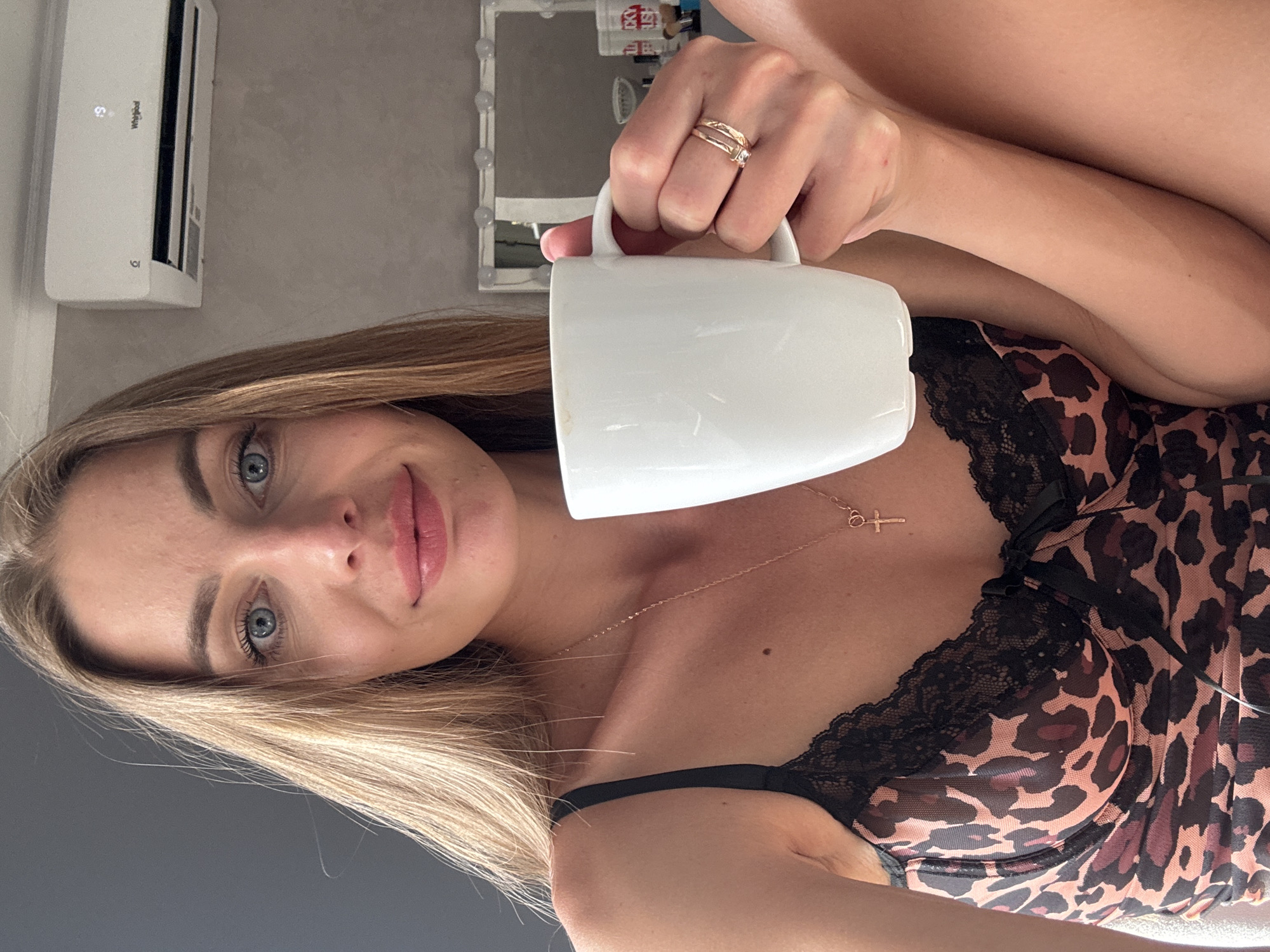 A woman with blonde hair and blue eyes, wearing a leopard-print bra, holds a white mug of coffee. She has on a cross necklace and rings on her fingers. The background shows an air conditioner mounted on a gray wall. A mirror with lights is visible in the distance. The scene appears to be indoors, potentially in a bedroom or dressing area. There is no indication of the location being in Sochi, Russia.