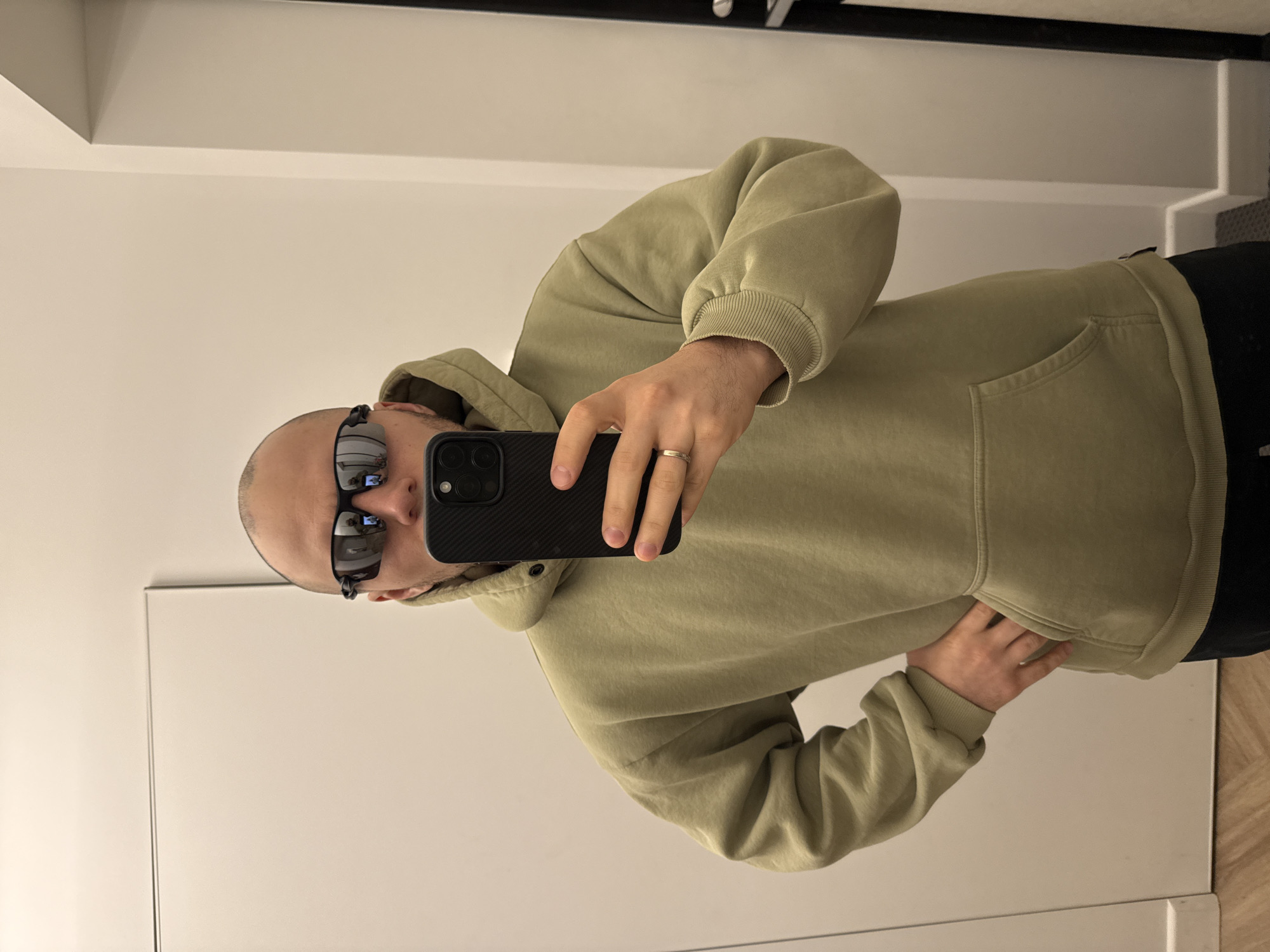 A bald man wearing sunglasses and a light green hoodie is taking a selfie in front of a mirror. He is holding a black iPhone in his right hand, and a wedding ring is visible on his left ring finger. The background consists of a white wall and a black metal bar. There is no indication of the location being in Krasnoyarsk, Russia.