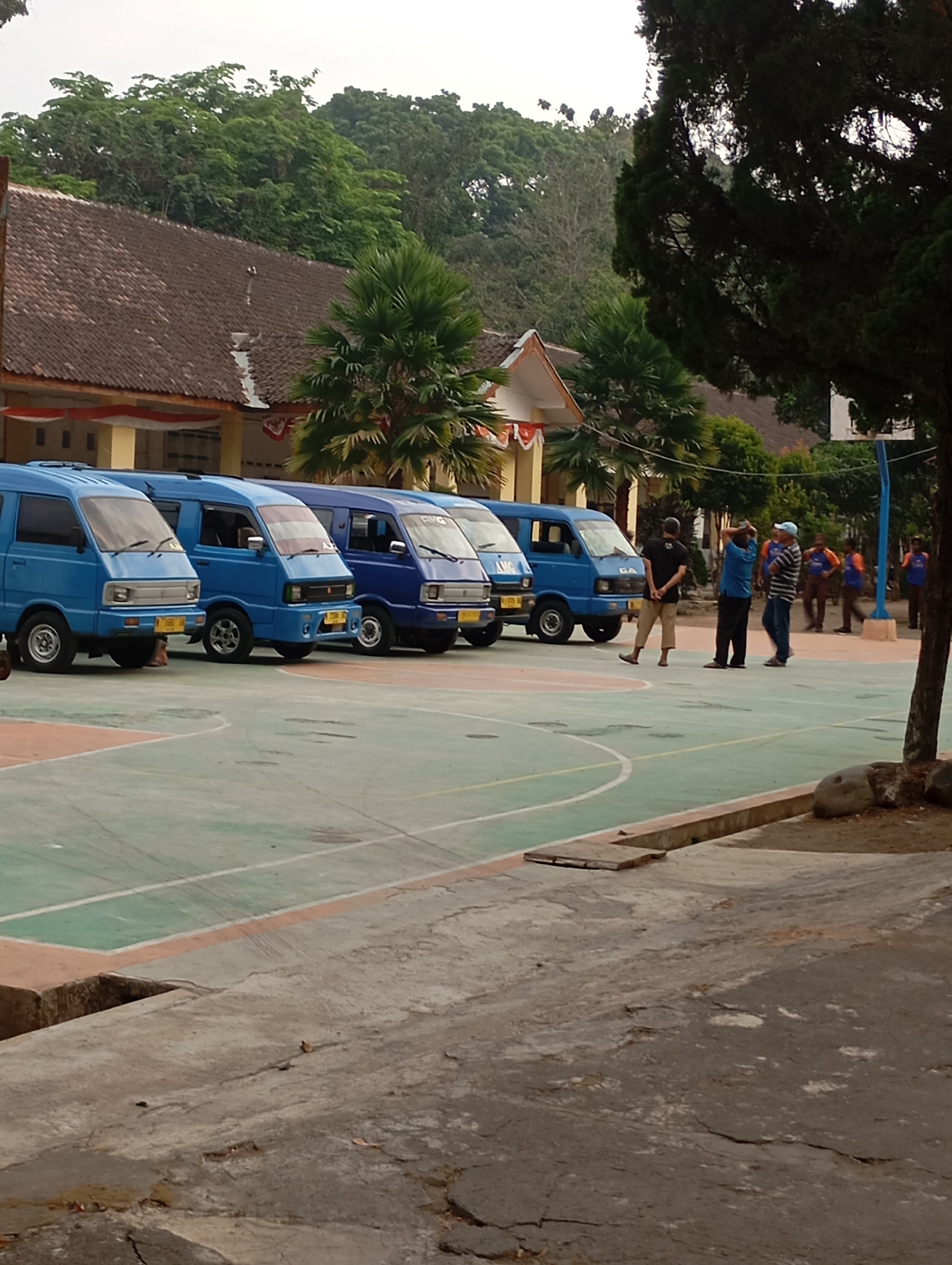 In Malang, Indonesia, this image captures an outdoor scene on what appears to be a school or sports complex. A line of several blue minivans is parked on a green and orange court. Three men are standing near the vans, seemingly in conversation. One man in a black shirt and tan pants stands with his back partially to the camera. Another man in a blue shirt and black pants is talking to a third man in a blue and white striped shirt and jeans. Further in the background, several other individuals are visible, some wearing orange shirts, standing near a basketball hoop. Behind the court, a low-lying building with a brown tiled roof and yellow walls is visible, decorated with red and white banners. Lush green trees and a large, dark green pine tree frame the scene. The foreground shows a cracked concrete surface, possibly a walkway or pathway. The sky is overcast.