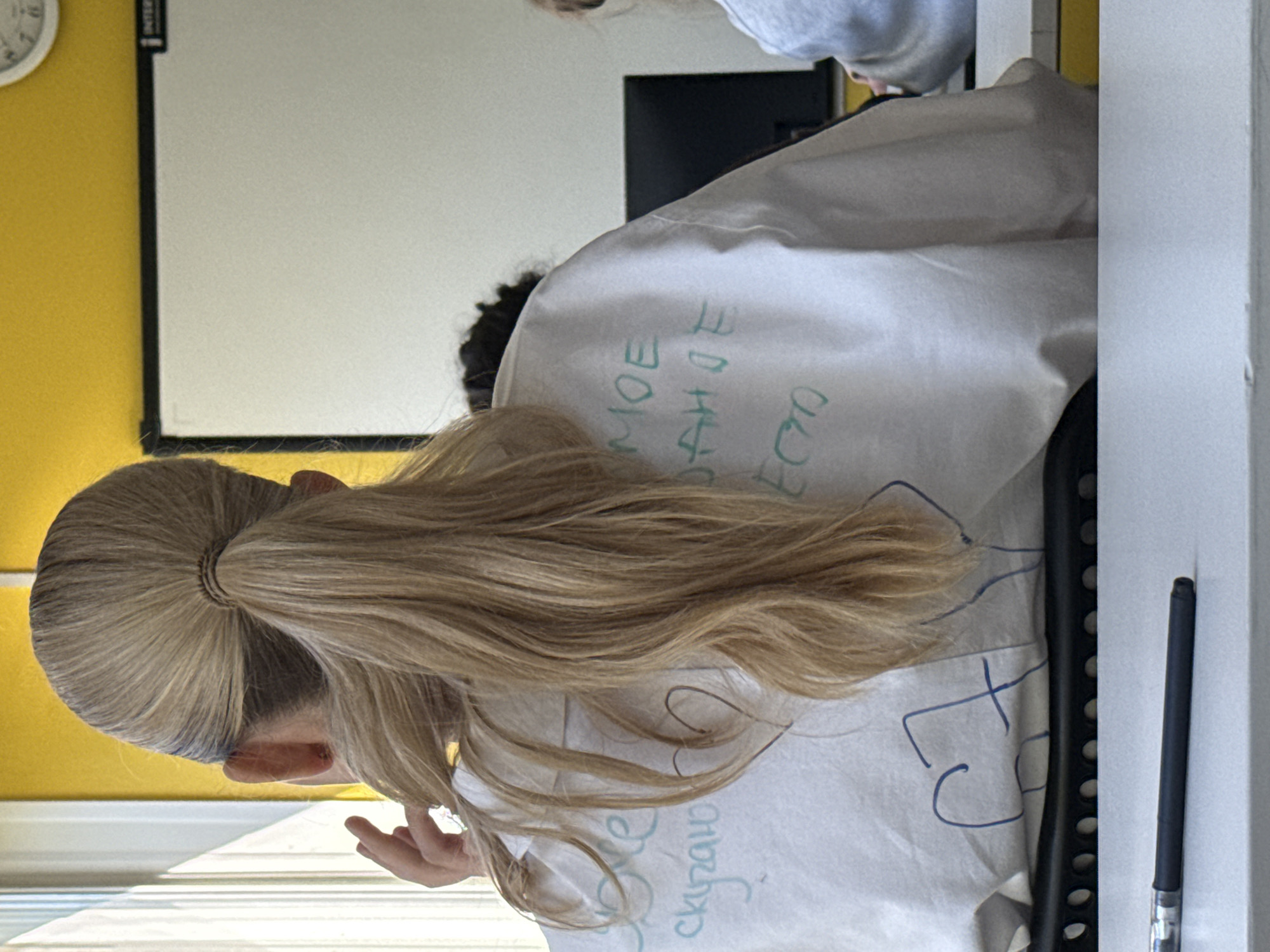 A blonde-haired person is seated in a classroom. They are wearing a white garment with Cyrillic writing on it, which includes the words "moe" and "danoe". The classroom has a yellow wall, a whiteboard, and a window. A pen rests on a desk or table near the person. The location is potentially in Krasnoyarsk, Russia, based on the Cyrillic writing.
