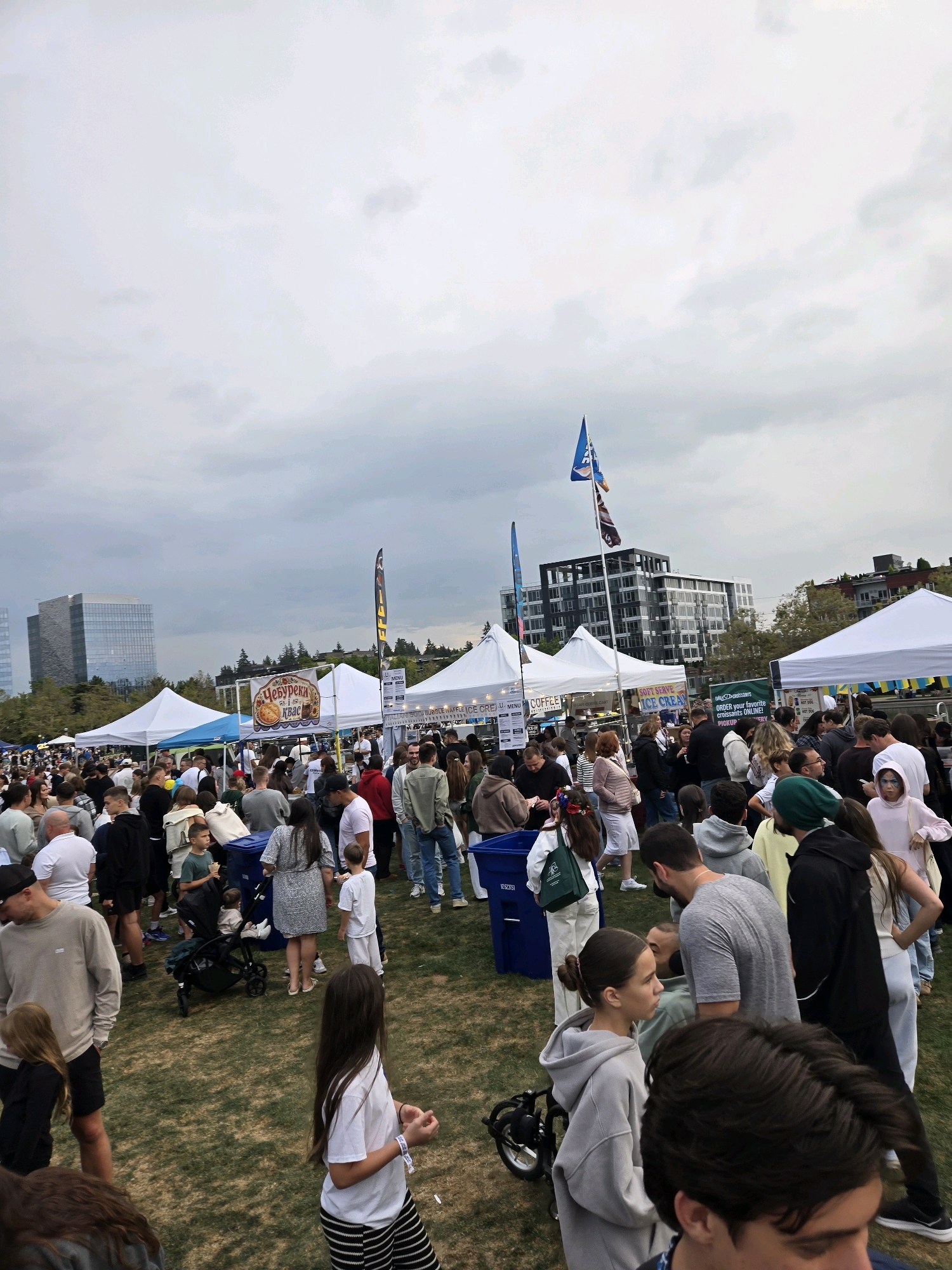 This is a wide-angle, eye-level photograph of an outdoor festival or market on a cloudy day. The scene is set in a grassy field with several white tents set up, suggesting food or merchandise vendors. In the background, modern glass-fronted buildings are visible, characteristic of the city of Bellevue, Washington, United States of America. A large crowd of people of various ages are milling about between the tents and in the open areas. Some individuals are walking, others are standing and talking, and a few appear to be browsing. There are strollers, trash receptacles, and promotional flags visible. The overall atmosphere suggests a community event.