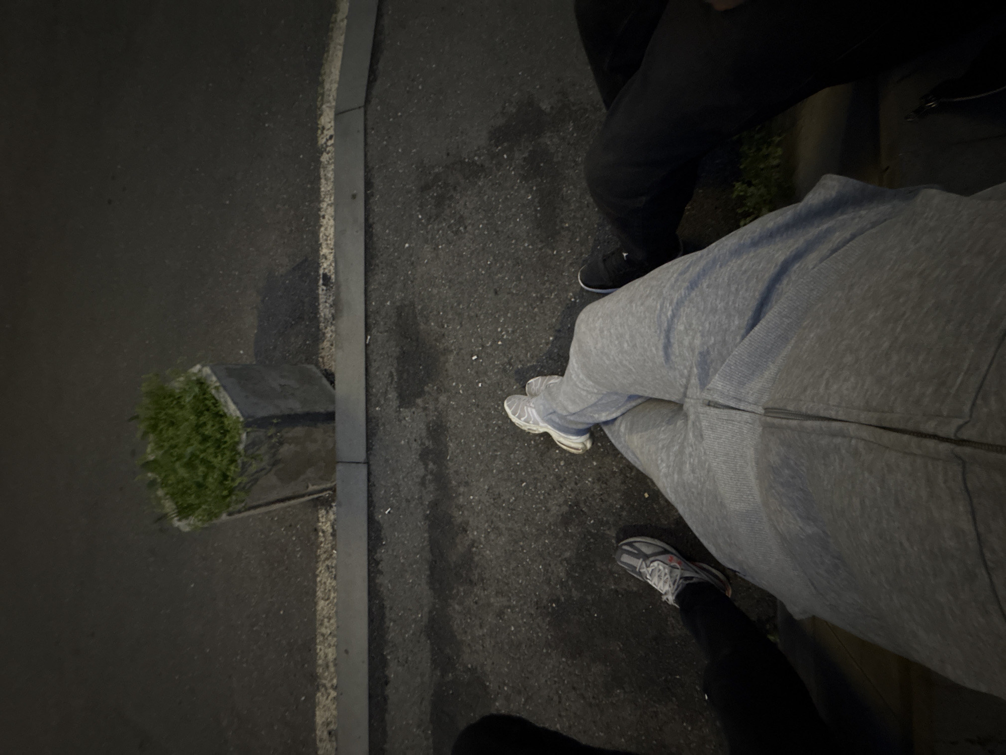 The image shows a street scene at night. A curb and a small concrete planter with grass are visible on the left. Two people are standing on the pavement. One person is wearing black pants and black shoes. The other person is wearing a gray tracksuit and white shoes. The location is identified as Yerevan, Armenia, but no other identifying information can be inferred from the picture. The camera seems to be pointed downwards from a height.