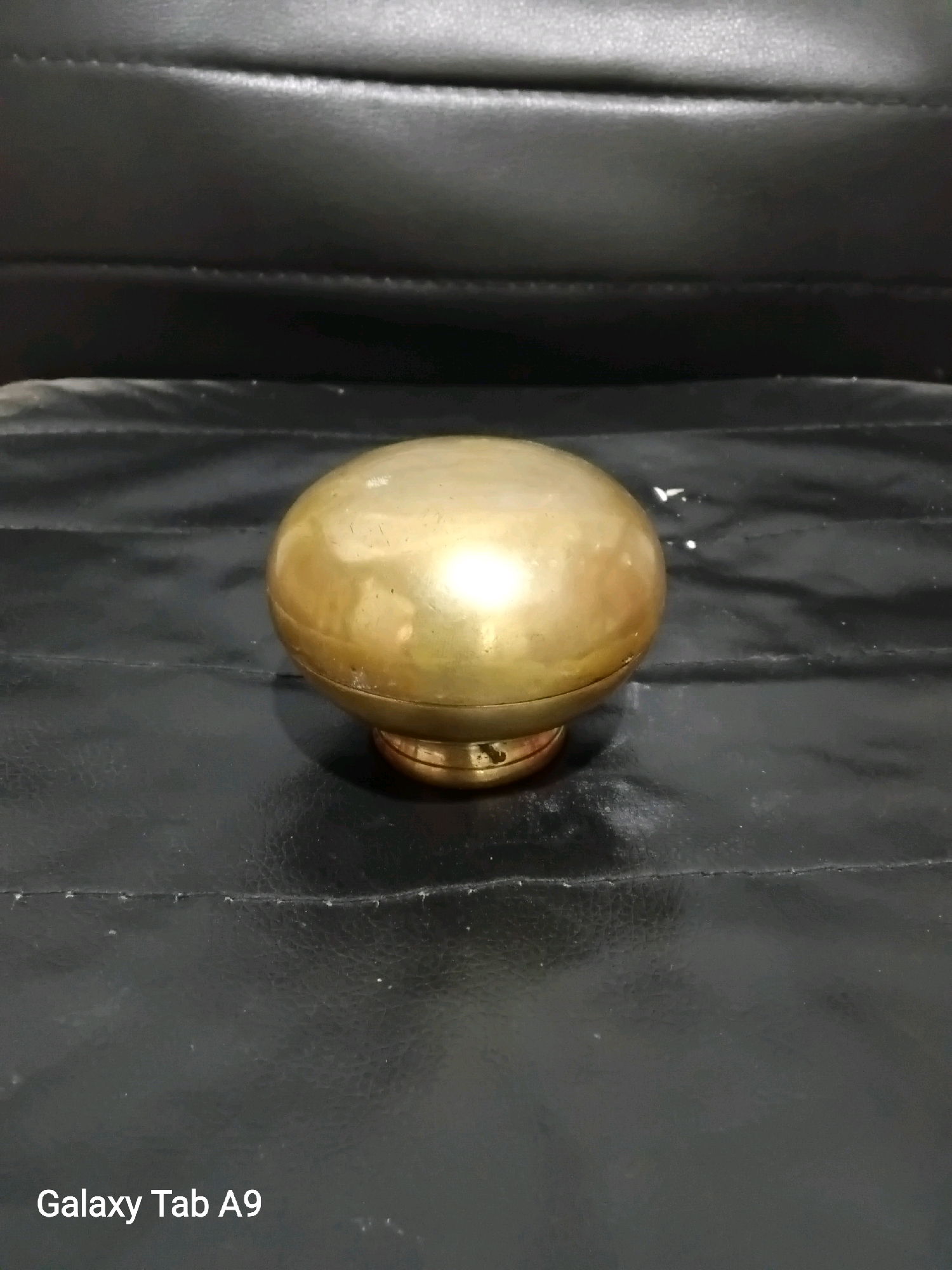A brass spherical object, possibly a drawer pull or decorative finial, rests on a black, textured surface. The object is round and has a metallic sheen, reflecting light from an unseen source. It is elevated on a small, circular base, also made of brass. The black surface appears to be some kind of upholstery, possibly leather, with visible stitching and creases. The lighting is dim, with the primary illumination focused on the brass object, creating highlights and shadows. The image is captured from a slightly elevated, eye-level perspective. The "Galaxy Tab A9" watermark is visible in the lower left corner, indicating the device used for photography and a potential location context of Karangan, Indonesia, if the metadata associated with the image were to be considered.