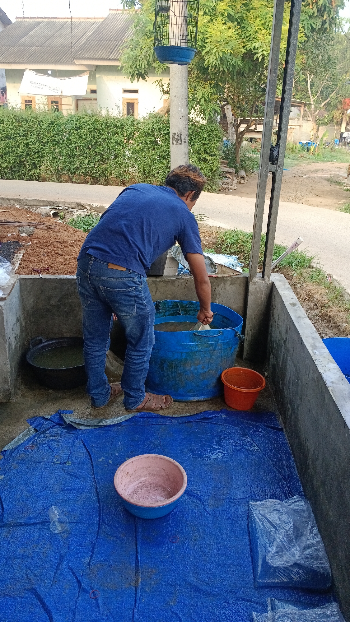 The image depicts a man standing in a shaded, outdoor area in Parung, Indonesia, engaged in an activity involving a large blue plastic tub. The man, wearing a blue t-shirt and blue jeans, is using a shovel to mix something within the tub, which appears to contain a mixture of water and possibly soil or feed. Next to the blue tub is a smaller orange plastic bucket. In the foreground, a blue tarp is spread out, with a pink and blue bowl and a clear plastic cup placed on it. To the left of the man, a black basin holds murky green liquid. The background reveals a residential setting with a house, a dense green hedge, and trees. A birdcage is suspended from a pole to the left of the man. The overall environment suggests a functional outdoor space, possibly for animal husbandry or gardening.