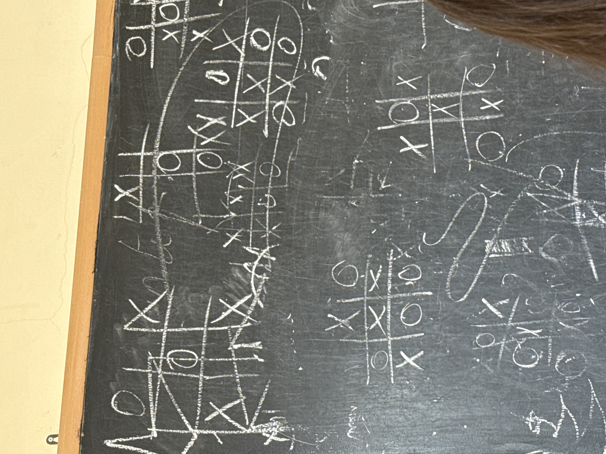 The image shows a chalkboard filled with numerous tic-tac-toe games drawn in white chalk. The games are in various states of completion, some with clear winners, others appearing unfinished or scribbled over. The chalkboard is framed with a wooden border and is attached to a wall, visible on the left side of the image. The wall is painted in a light, creamy color. The overall environment appears to be a classroom or similar educational setting. There is no information on this photo that tells us about location or city of Yerevan, Armenia.