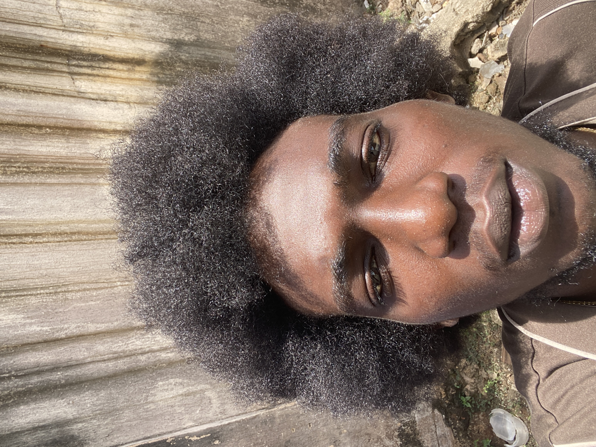 The image shows a close-up of a person with dark skin and an afro hairstyle. The person is wearing a brown outfit with a white stripe. The background consists of a weathered, striated surface and some dirt with small green plants. While it's impossible to determine the exact location without more information, the environment suggests an outdoor setting, potentially in a residential or less-developed area. Lagos, Nigeria, is speculated as the location.
