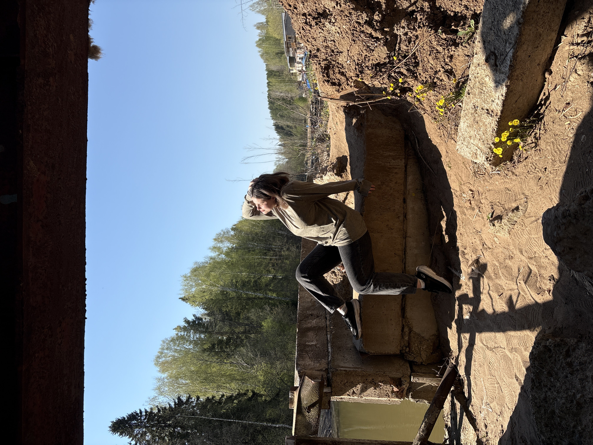 A woman is pictured in what appears to be an abandoned or dilapidated structure. She is wearing dark pants, a light-colored long-sleeved shirt, and sneakers. She has her hand up to her forehead, perhaps shielding her eyes. The setting includes dirt, vegetation including yellow flowers, and remnants of concrete. In the background, there is a forest and possibly some buildings. The image's context suggests a location near Chaikovsky, Russia, due to the forest in the background and the image's provenance.