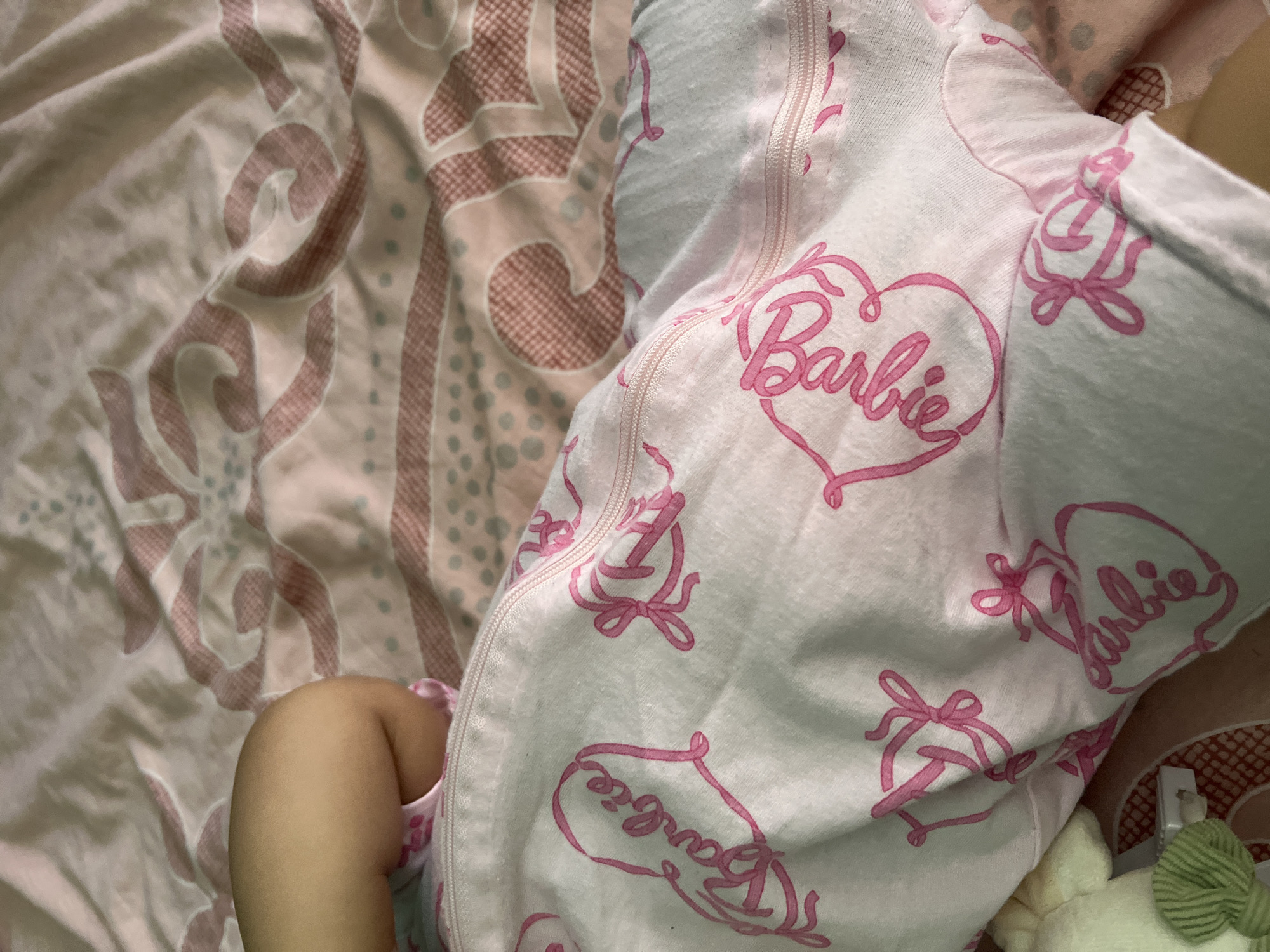 A close-up, low-angle shot shows a baby's thigh and lower leg wrapped in a pink and white Barbie-themed swaddle. The swaddle has a zipper closure and is patterned with hearts and the word "Barbie" in cursive. The baby is lying on a pink and white patterned bedspread. The image is taken indoors, with no specific location in Plovdiv, Bulgaria identifiable.