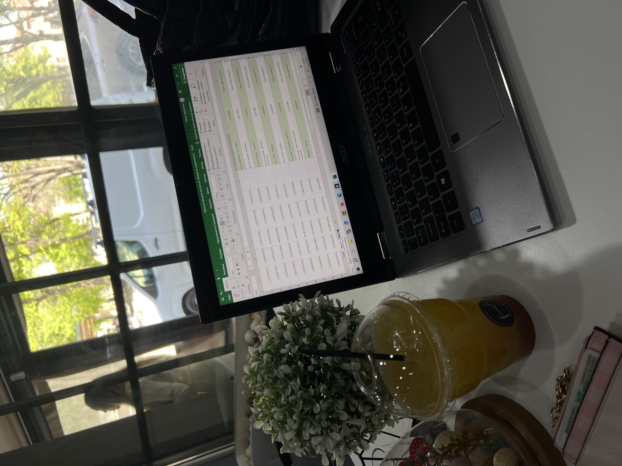 The image displays an indoor scene, likely a cafe or office in Plovdiv, Bulgaria, with a laptop open on a light-colored table next to a window. The central subject is an Acer laptop displaying a Microsoft Excel spreadsheet titled "Публични финанси паї поvo.xls" (Public finances). The spreadsheet contains columns of numbers and text in Bulgarian, including financial terms such as "Ръст на БВП" (GDP growth) and "Ръст на ЕКВ". The laptop's taskbar shows the time "11:30" and date "23.4.2026", along with a notification stating "Rainy days ahead". The keyboard has both Latin and Cyrillic characters, corresponding to the Bulgarian language. An Intel Core i5 sticker is visible on the laptop's palm rest.
To the right of the laptop, a clear plastic cup filled with a yellow-orange beverage and a black straw is present, next to a small artificial plant with green and white leaves. Partial views of decorative items under a glass dome and a pink and white checkered accessory, possibly a wallet or small bag, are also seen.
Through the window, an outdoor view shows vibrant green trees and part of a white vehicle, indicating a bright and clear day outside. A reflection of a person with long hair is subtly visible in the windowpane. The scene suggests a work or study session in a relaxed, well-lit environment.