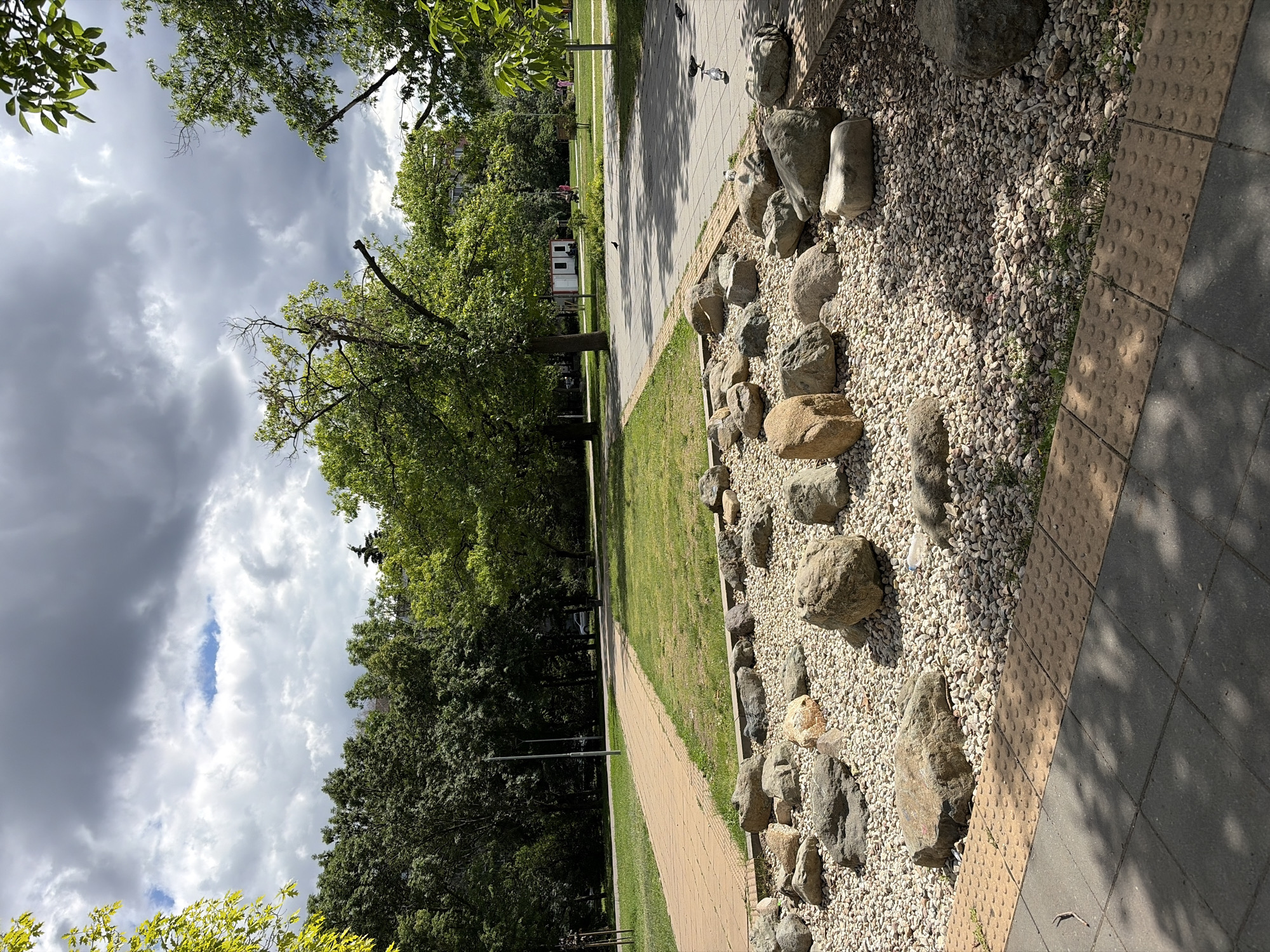 An outdoor shot captures a park on a cloudy day. In the foreground, a decorative arrangement of large rocks is set within a bed of small pebbles. To the left, a paved walkway runs adjacent to a grassy area. Further back, a dense line of trees forms a backdrop, with a few buildings visible through the foliage. Several birds are present on a paved path in the middle ground. The sky is heavily overcast, with patches of blue sky visible through the clouds.